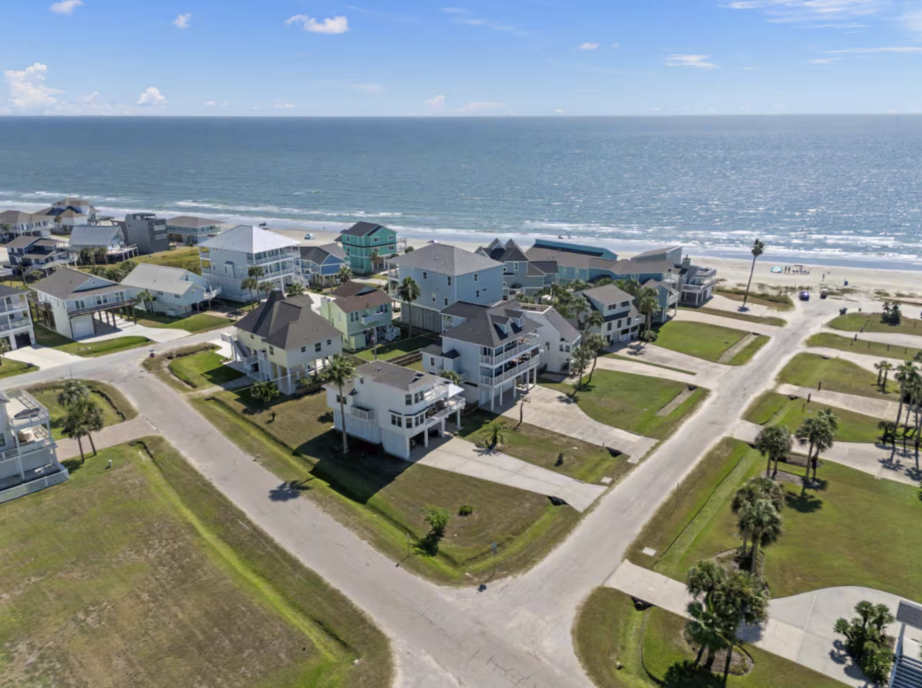 Aerial view of a beachside neighborhood with multicolored houses, green lawns, palm trees, and a sandy beach with the ocean in the background under a partly cloudy sky.