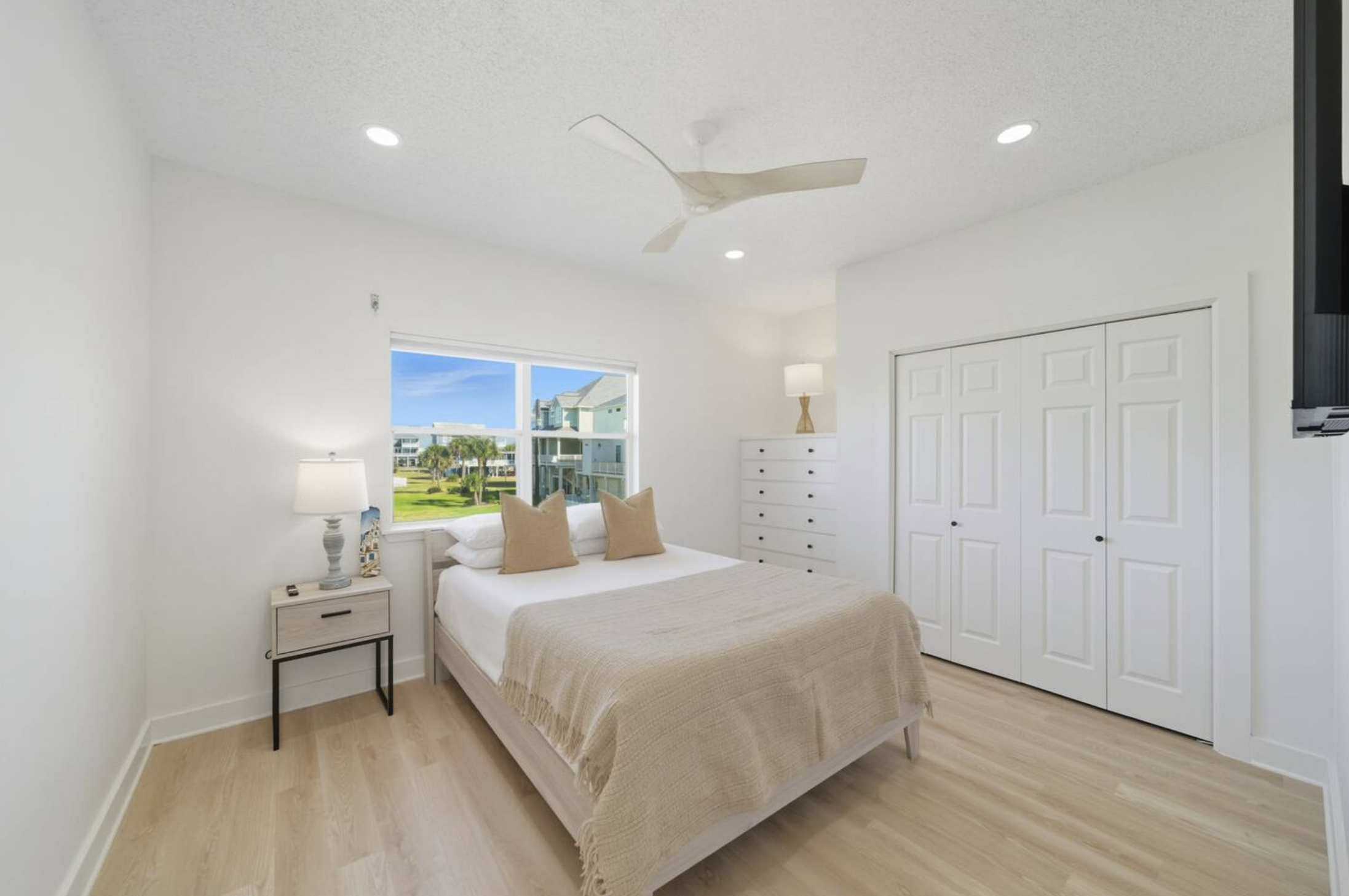 Bright bedroom with white walls, a bed with beige bedding and pillows, a nightstand with a lamp, a window showing a view of colorful buildings and palm trees, a white dresser, and a closet with double doors.