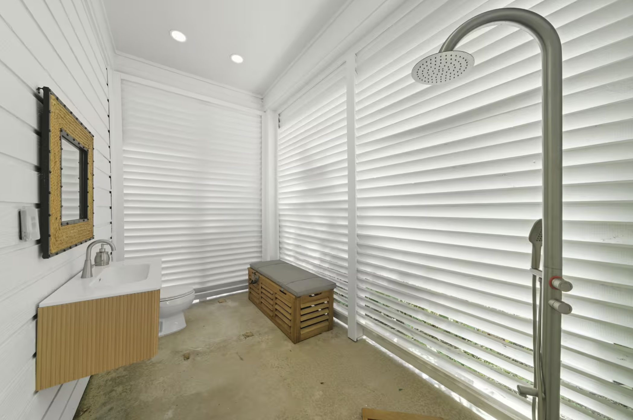 An outdoor shower area with a rain shower head, a small sink with a wooden cabinet, a bench with a cushion, and white horizontal siding walls with shutters.