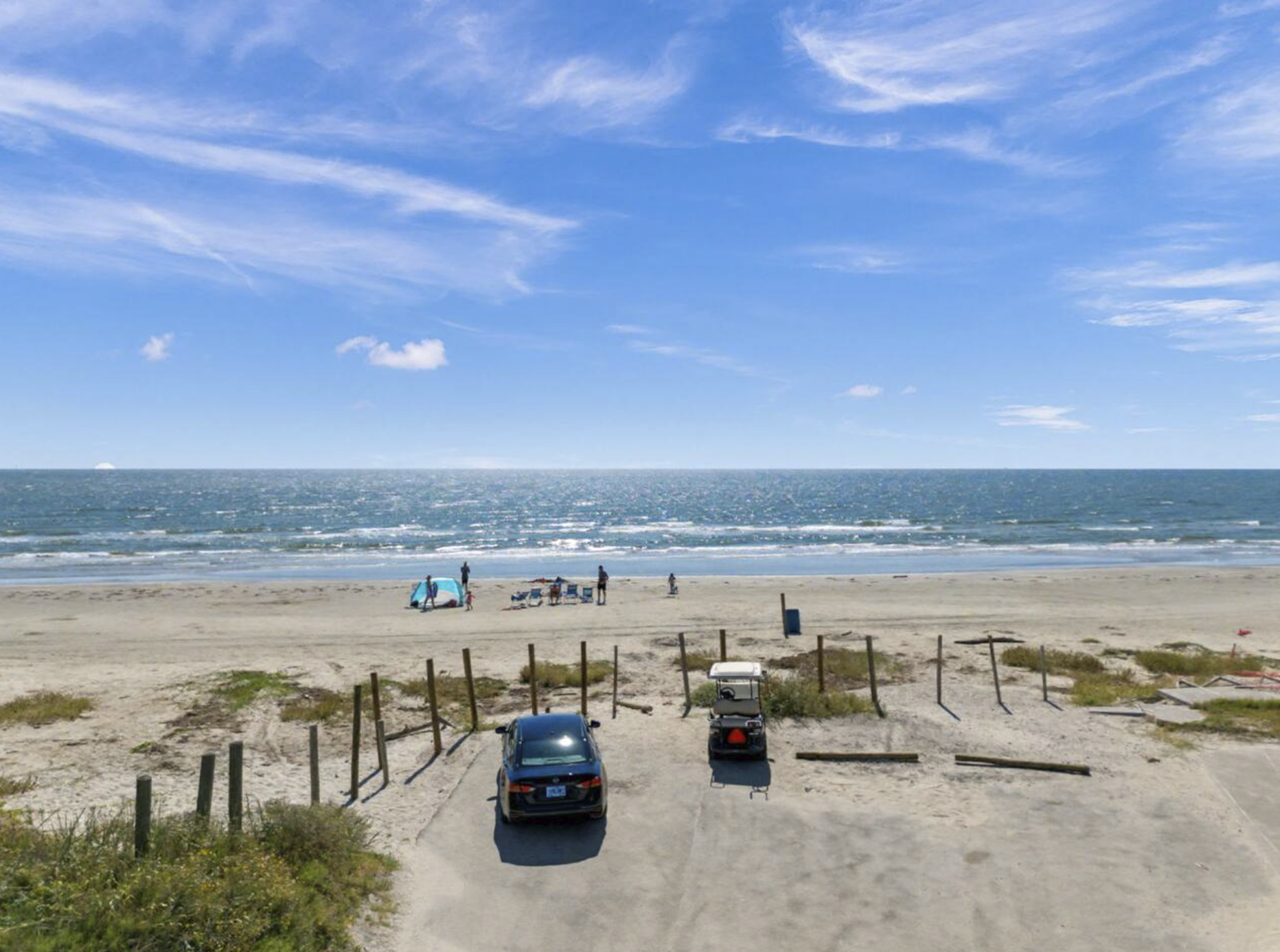 Beach scene with cars parked in the foreground, sandy dunes with some greenery, and a few people near a tent and chairs on the beach. The ocean with gentle waves and a partly cloudy blue sky are in the background.