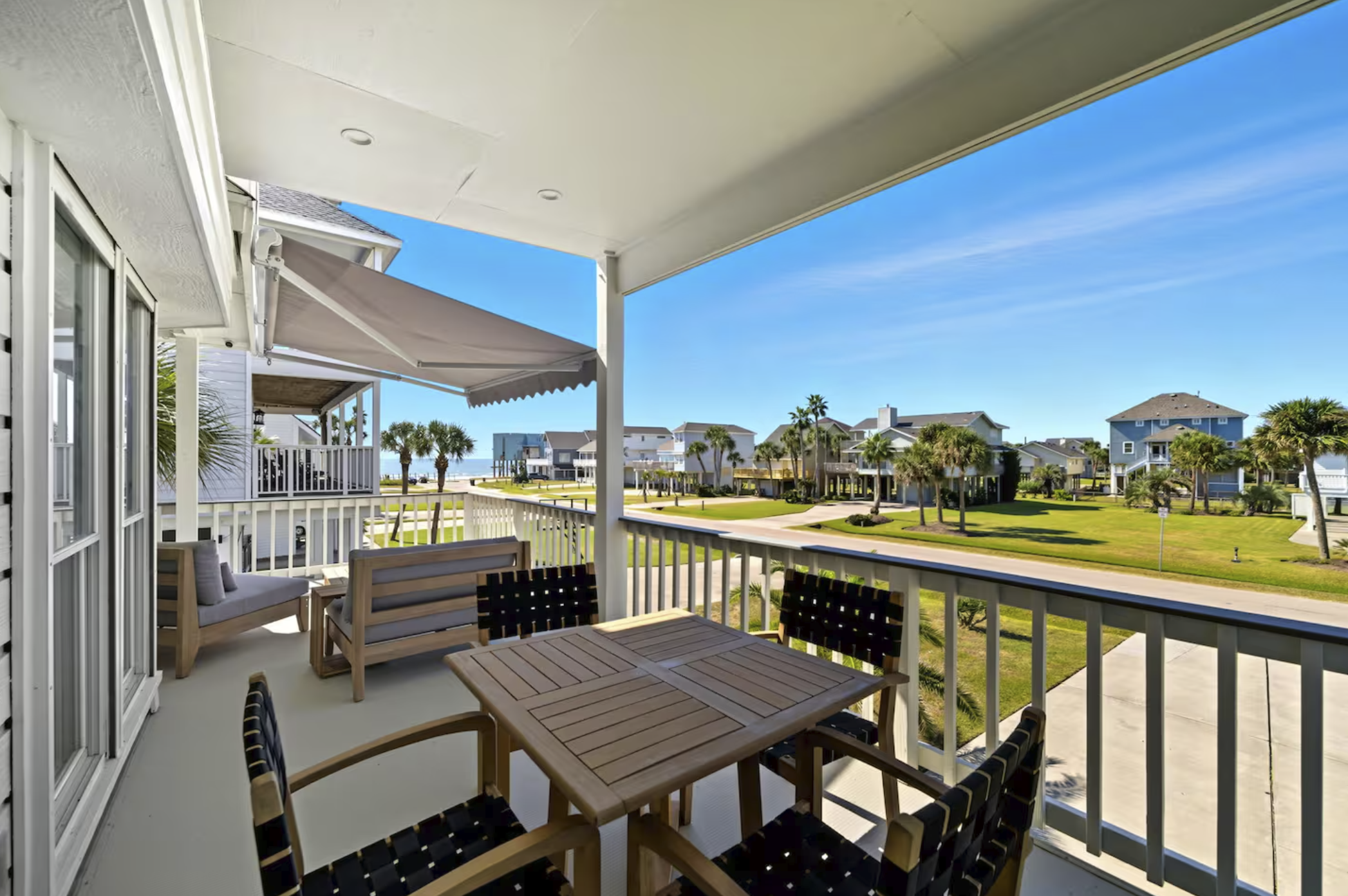 Balcony with outdoor furniture overlooking a neighborhood of beach houses with palm trees, under a clear blue sky.