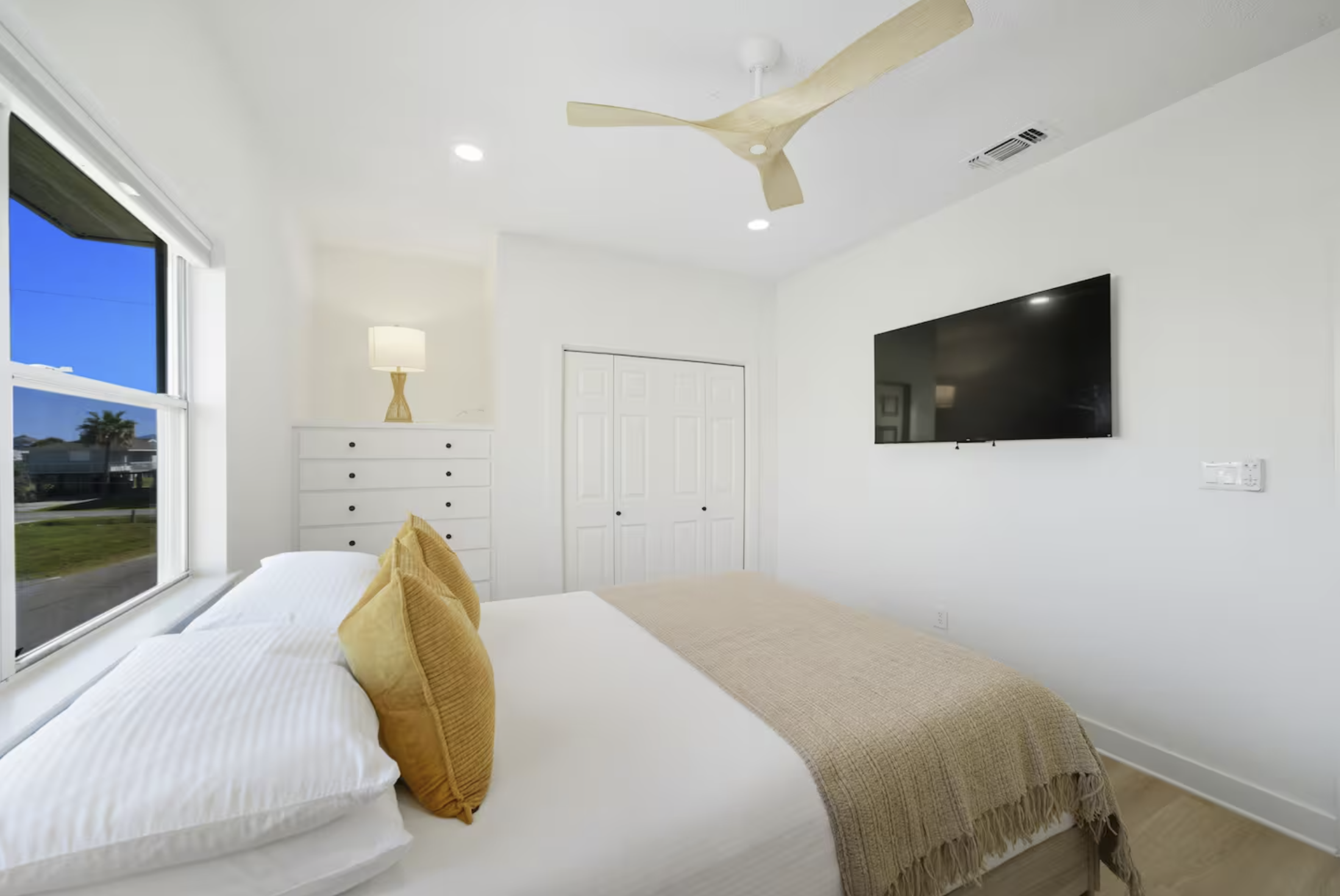 Cozy modern bedroom with white walls, bed with white sheets and beige blanket, two yellow pillows, a window showing a blue sky outside, a TV on the wall, a white dresser with a small lamp, ceiling fan, and closet doors.