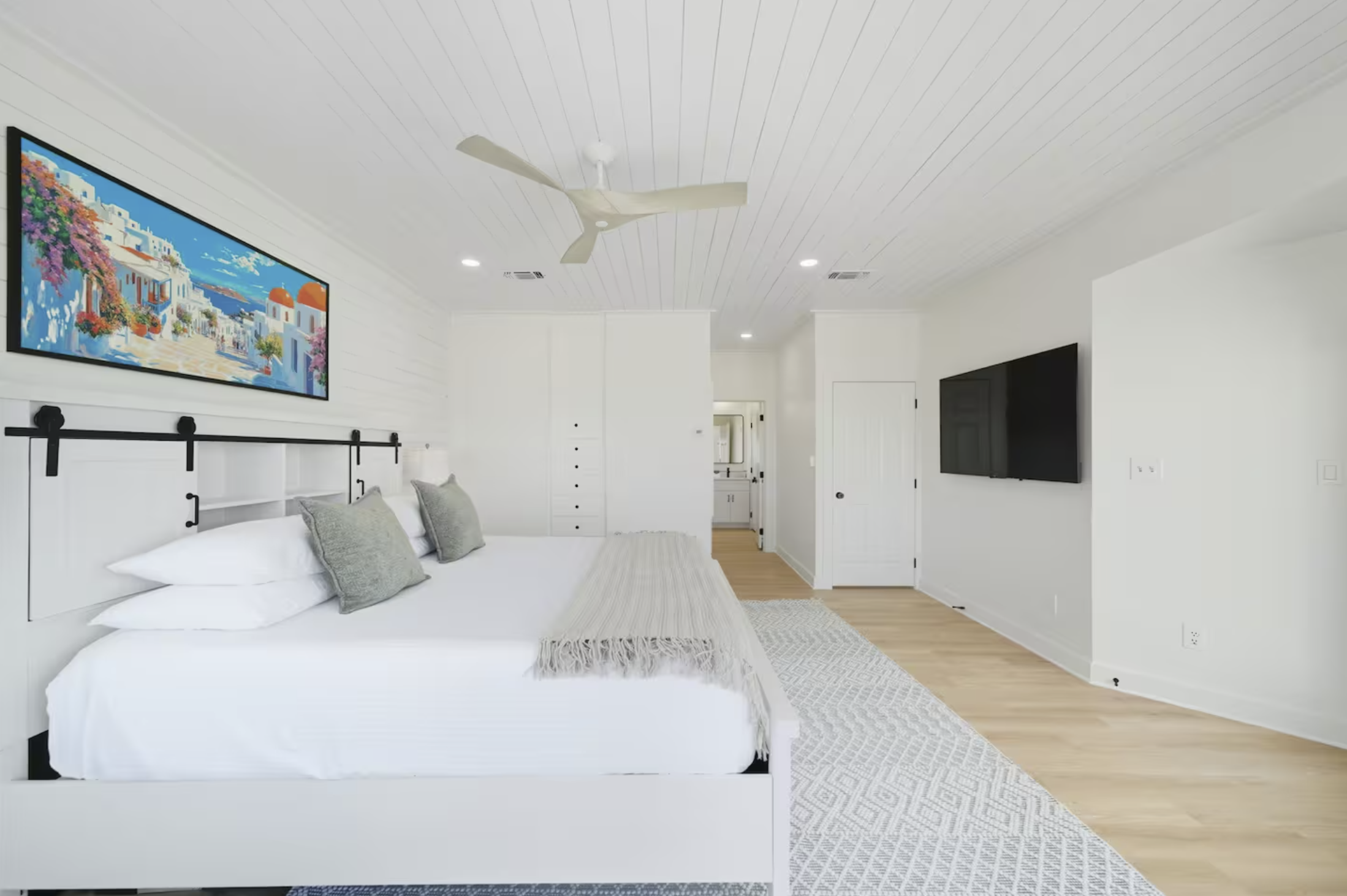 Bright modern bedroom with white walls and ceiling, featuring a large bed with white linens and multiple pillows, a hanging black metal headboard, a patterned area rug, a flat-screen TV on the wall, a ceiling fan, and a door leading to a bathroom wit