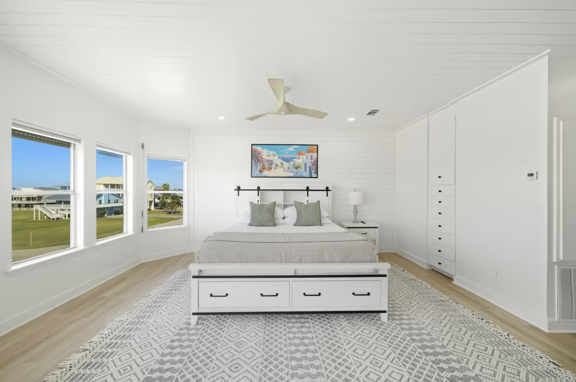 Bright bedroom with white walls, wooden floor, large windows, a bed with gray pillows, a white bed frame with drawers, a nightstand with a lamp, and a colorful artwork above the bed.