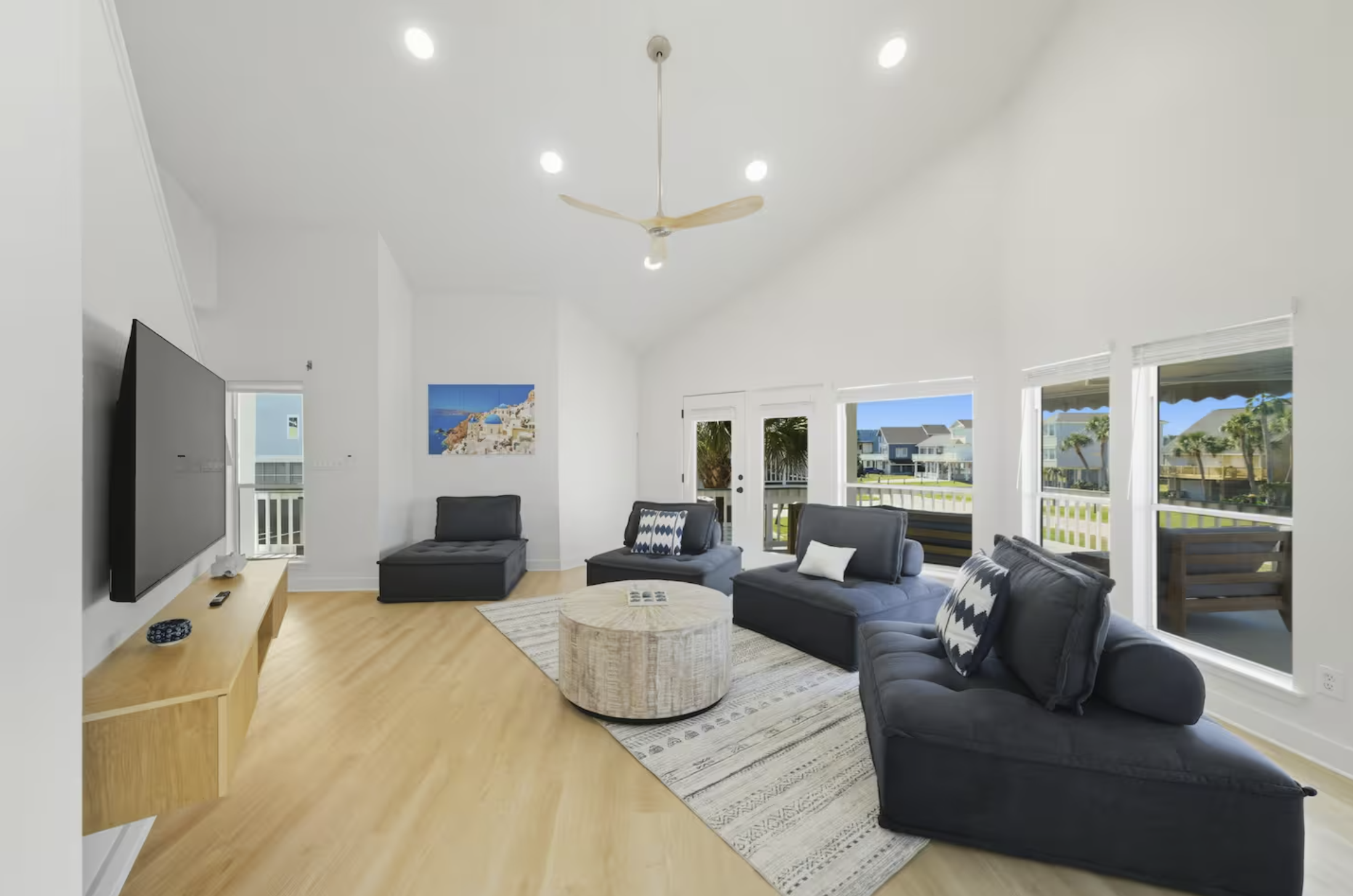 Bright living room with black sofas, a round beige ottoman, large windows, and a ceiling fan.