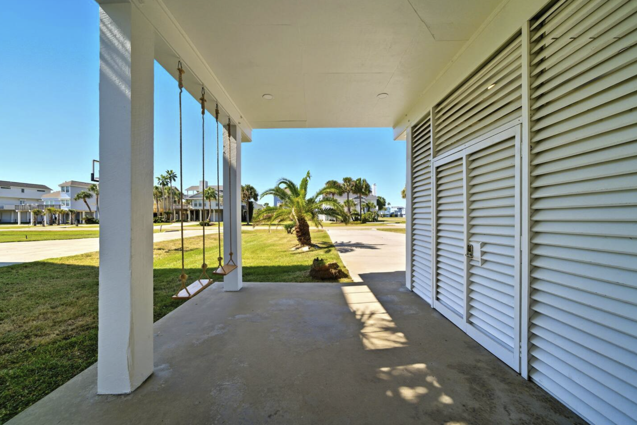 Front porch of a house with two wooden swings hanging from the ceiling, a white side wall with vented shutters, a grassy yard with palm trees, neighboring houses, and a bright blue sky.