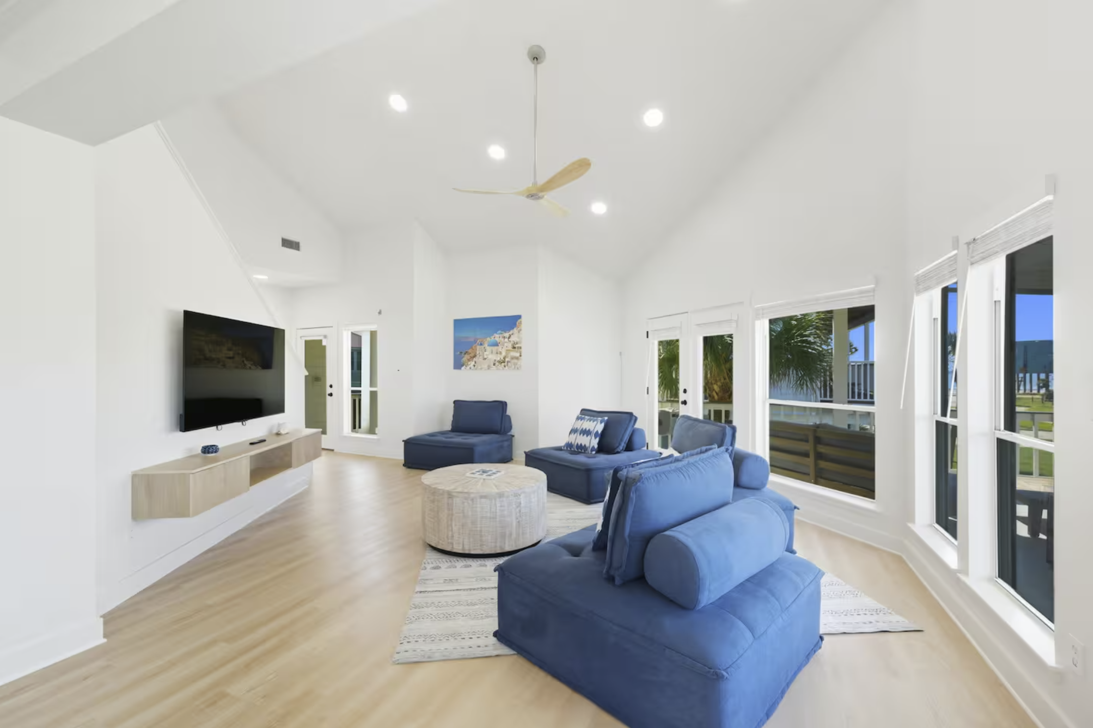 Bright living room with white walls, light wood floor, blue sofas, wall-mounted TV, large windows, glass doors leading outside, ceiling fan, wall art, and a small patterned rug.