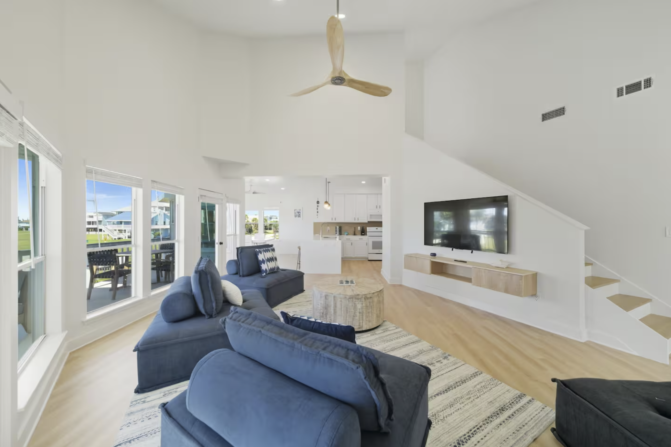 Living room with blue sofas, a beige round coffee table, a wall-mounted TV, large windows showing a porch with outdoor furniture, open kitchen, and ceiling fan.