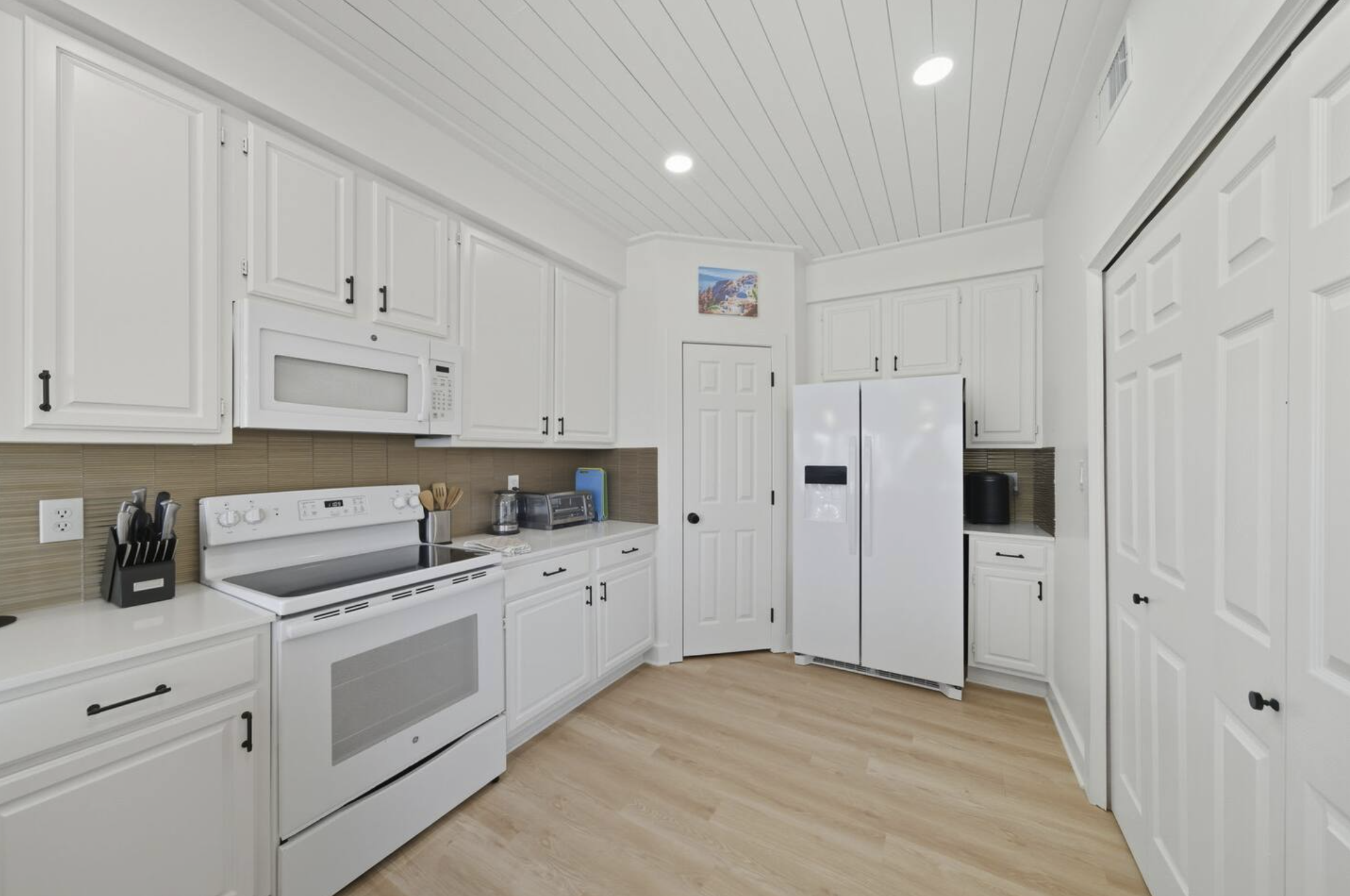 A bright kitchen with white cabinets, a white stove, microwave, and refrigerator, beige backsplash, and light wood floors.