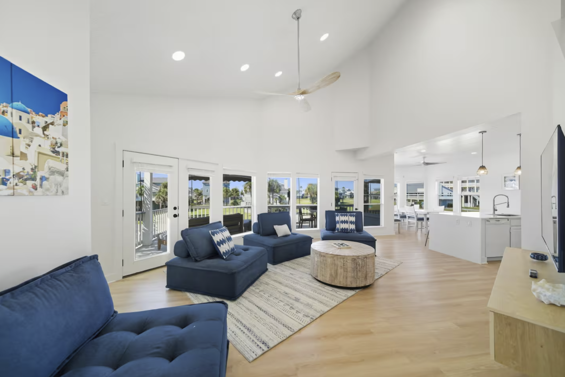 Bright living room with blue couches, large windows, and a view of a porch and outdoor area, with a white kitchen and dining space in the background.