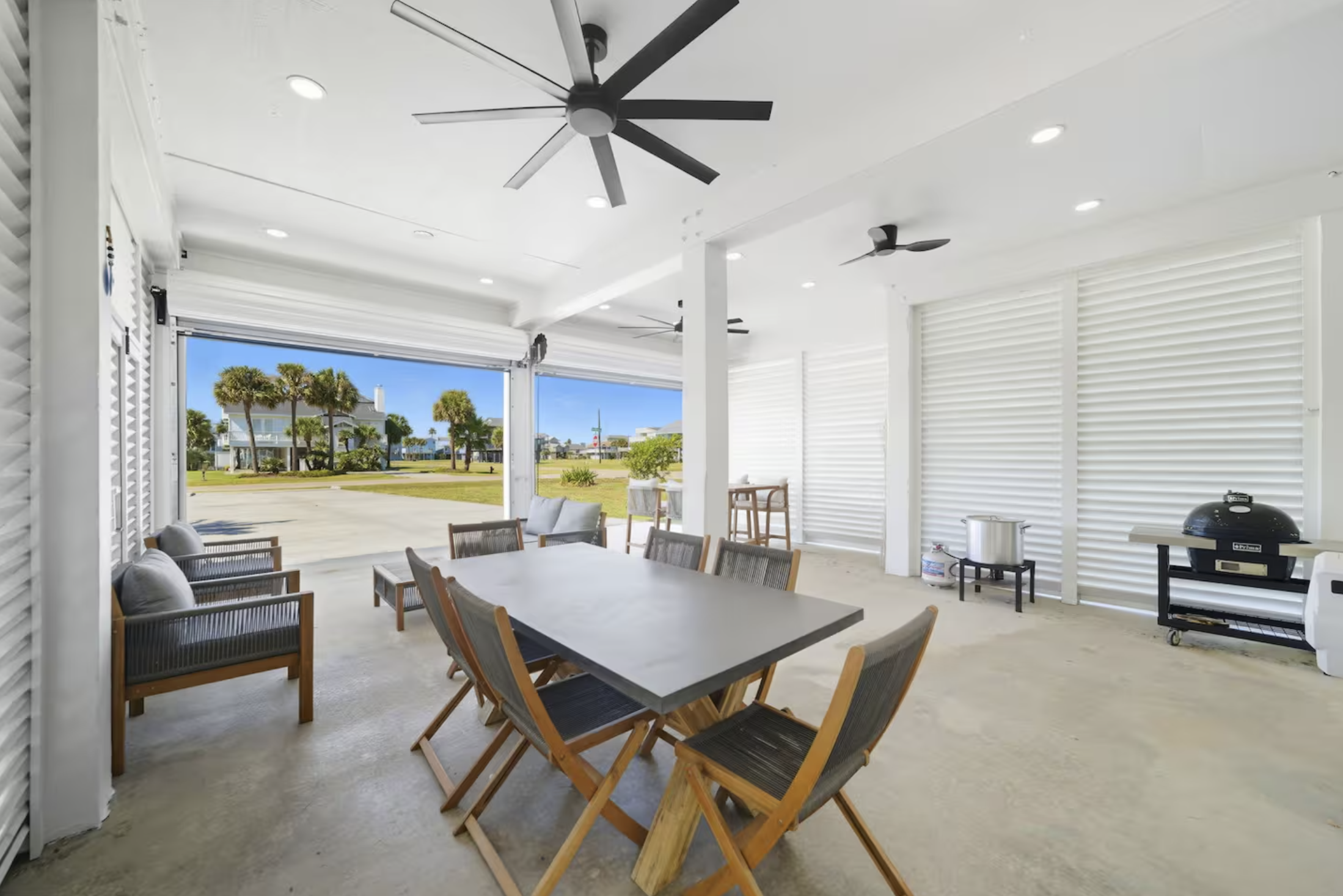 Outdoor covered patio area with white walls and ceiling, ceiling fans, a large open garage door revealing a view of a grassy lawn with palm trees and houses, a dining table with six chairs, a black grill, and various outdoor cooking equipment.