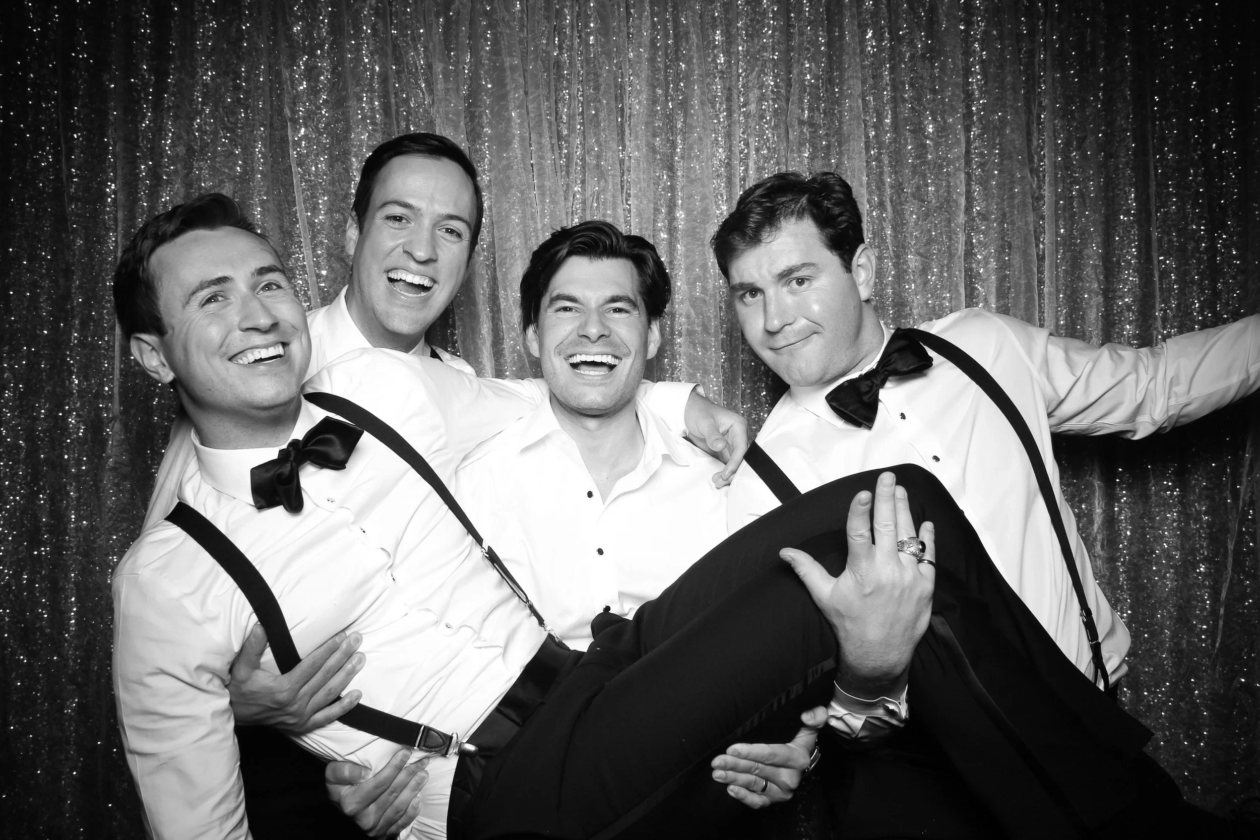 Black and white photo of five men dressed in formal attire, with three of them wearing suspenders and bow ties, celebrating together in front of a glittering backdrop. One man is being held horizontally by the others, all smiling and having a good time.