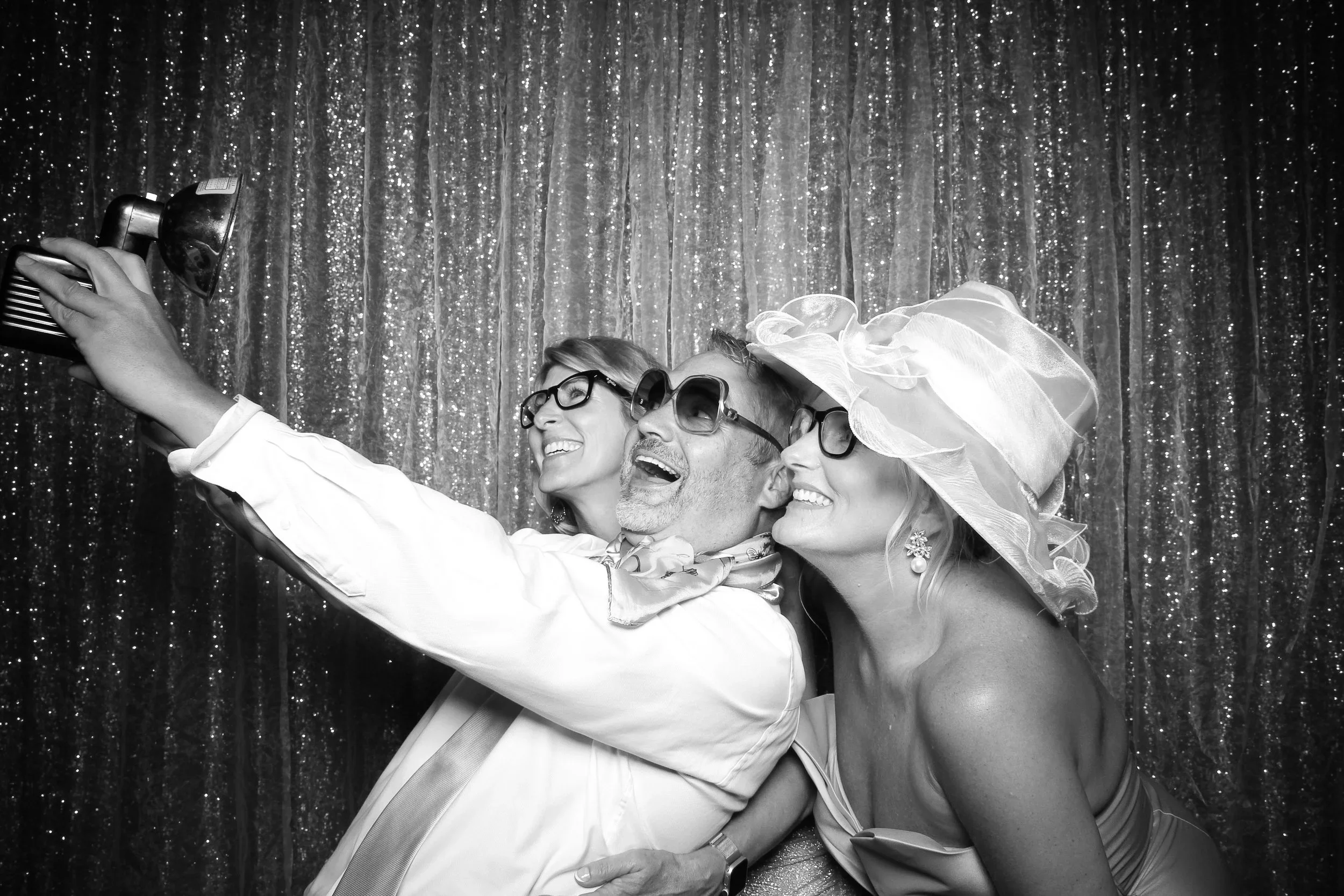 Three people smiling and taking a selfie in front of a sparkly backdrop. The man in the middle is wearing sunglasses and a scarf, and the women on either side are wearing large hats and glasses.