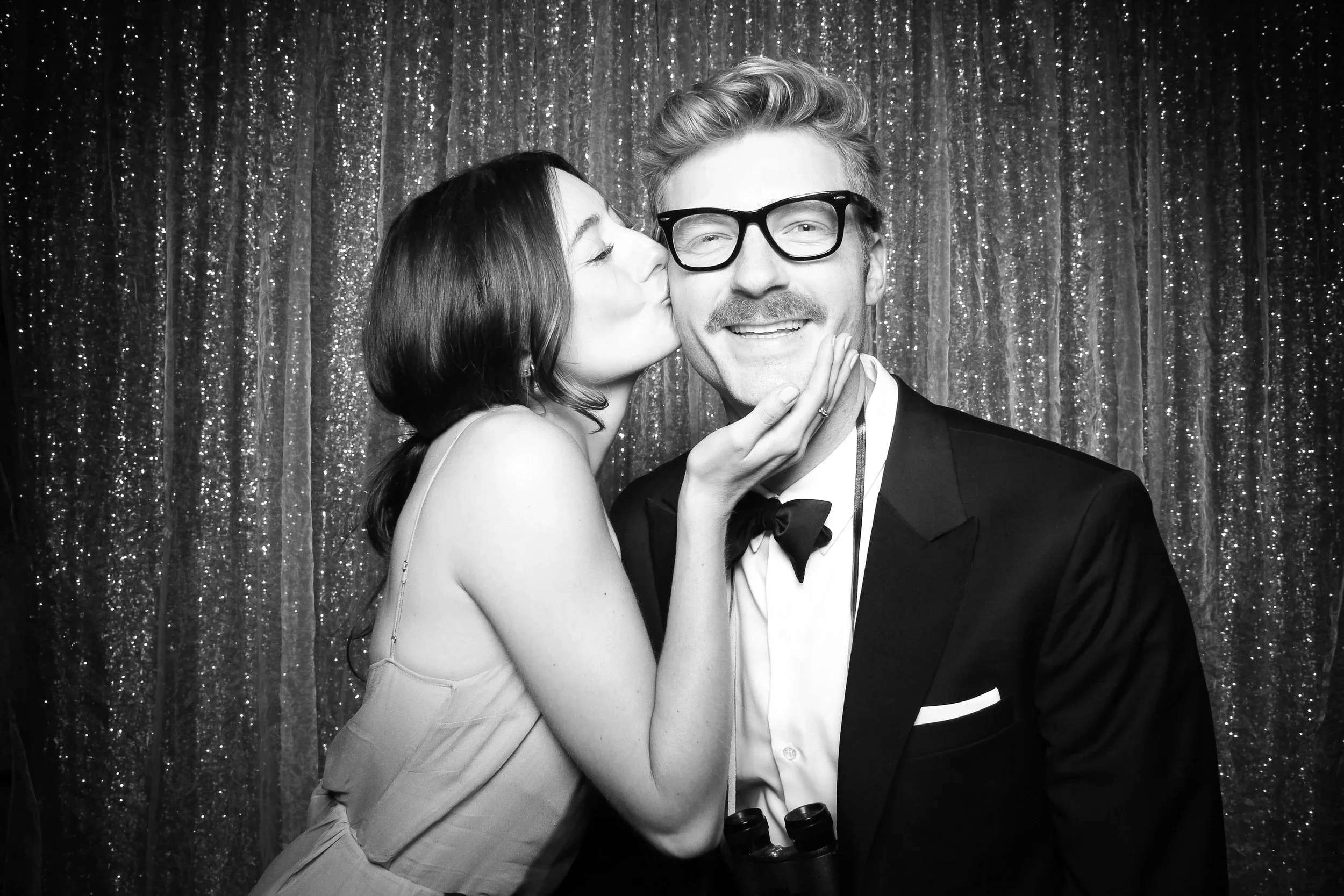 A woman kisses a man on the cheek at a formal event. The man is wearing a tuxedo with a bowtie and glasses, and they are in front of a sparkly curtain background.
