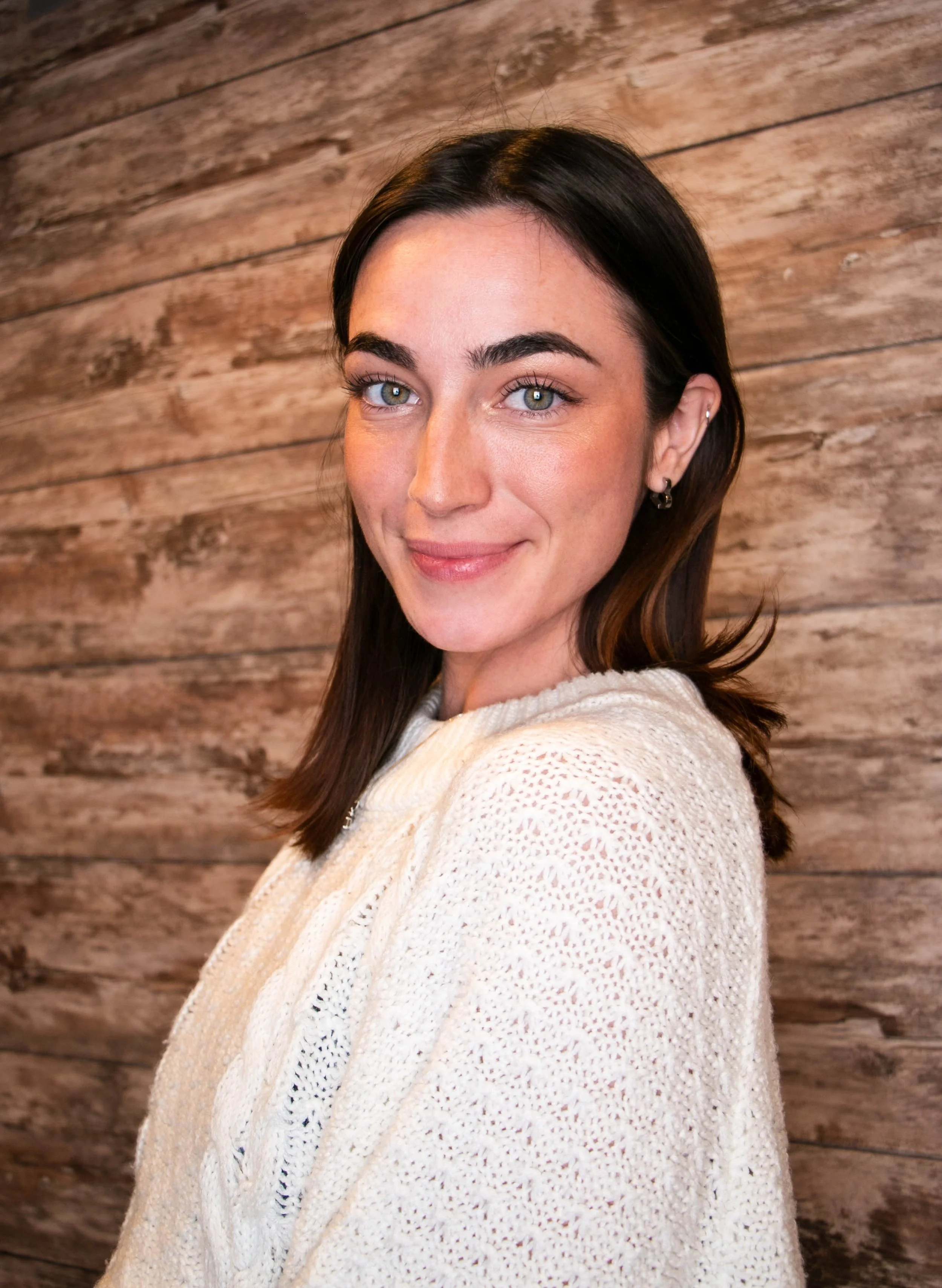 A young woman with dark hair and blue eyes smiling at the camera, wearing a white knitted sweater, standing in front of a wooden wall.