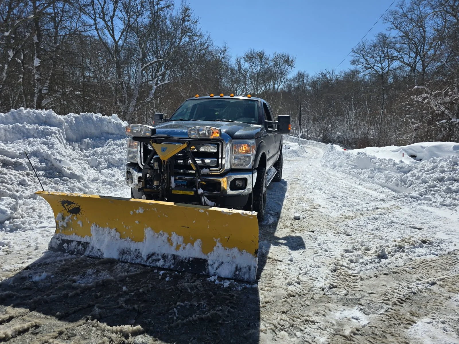A black pickup truck equipped with a yellow snowplow attached to the front is clearing snow from a rural snowy road surrounded by snow piles and leafless trees under a blue sky.