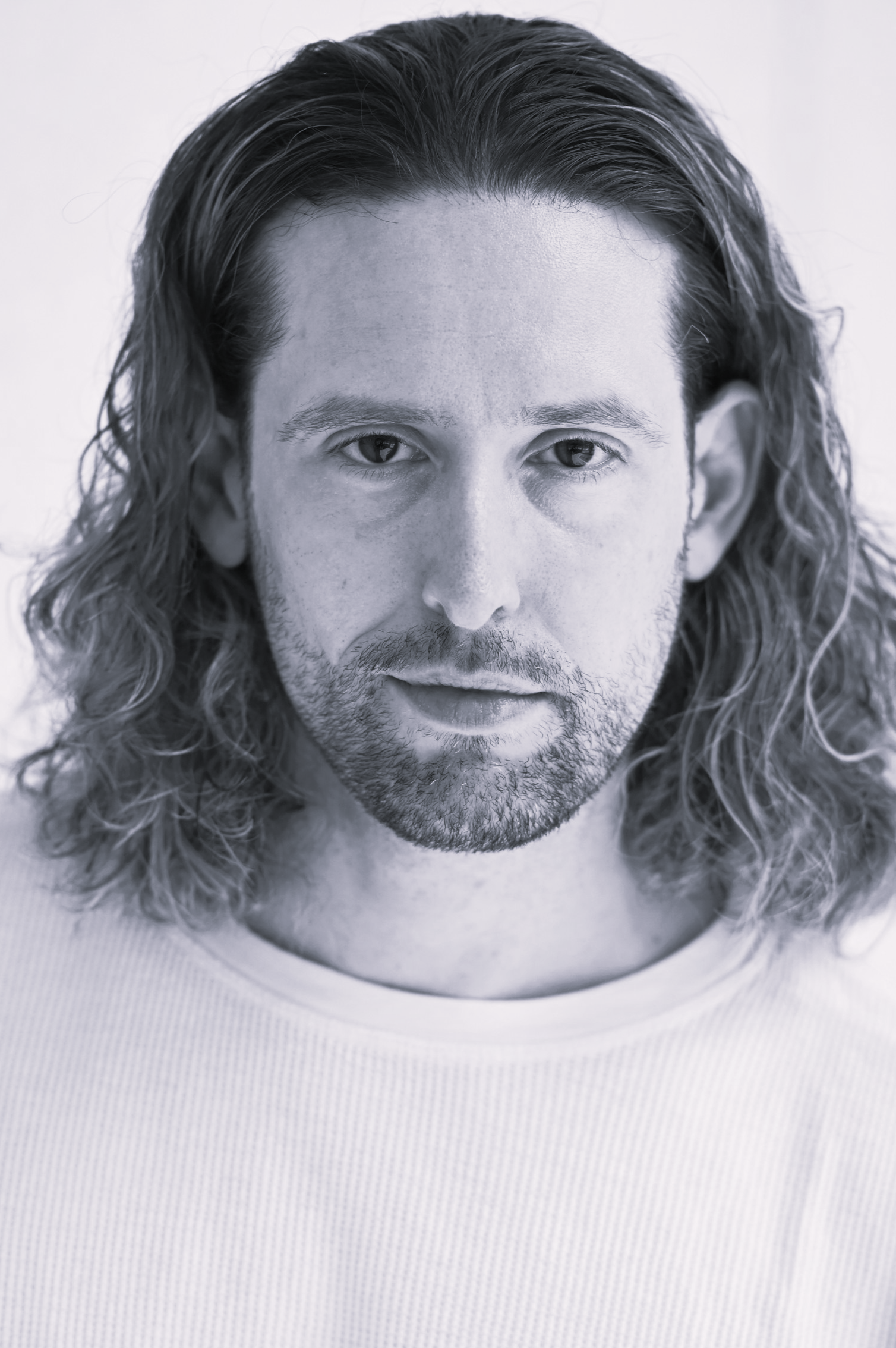 A black and white image of a young man with wavy hair in a white shirt looking at the camera with a serious expression.
