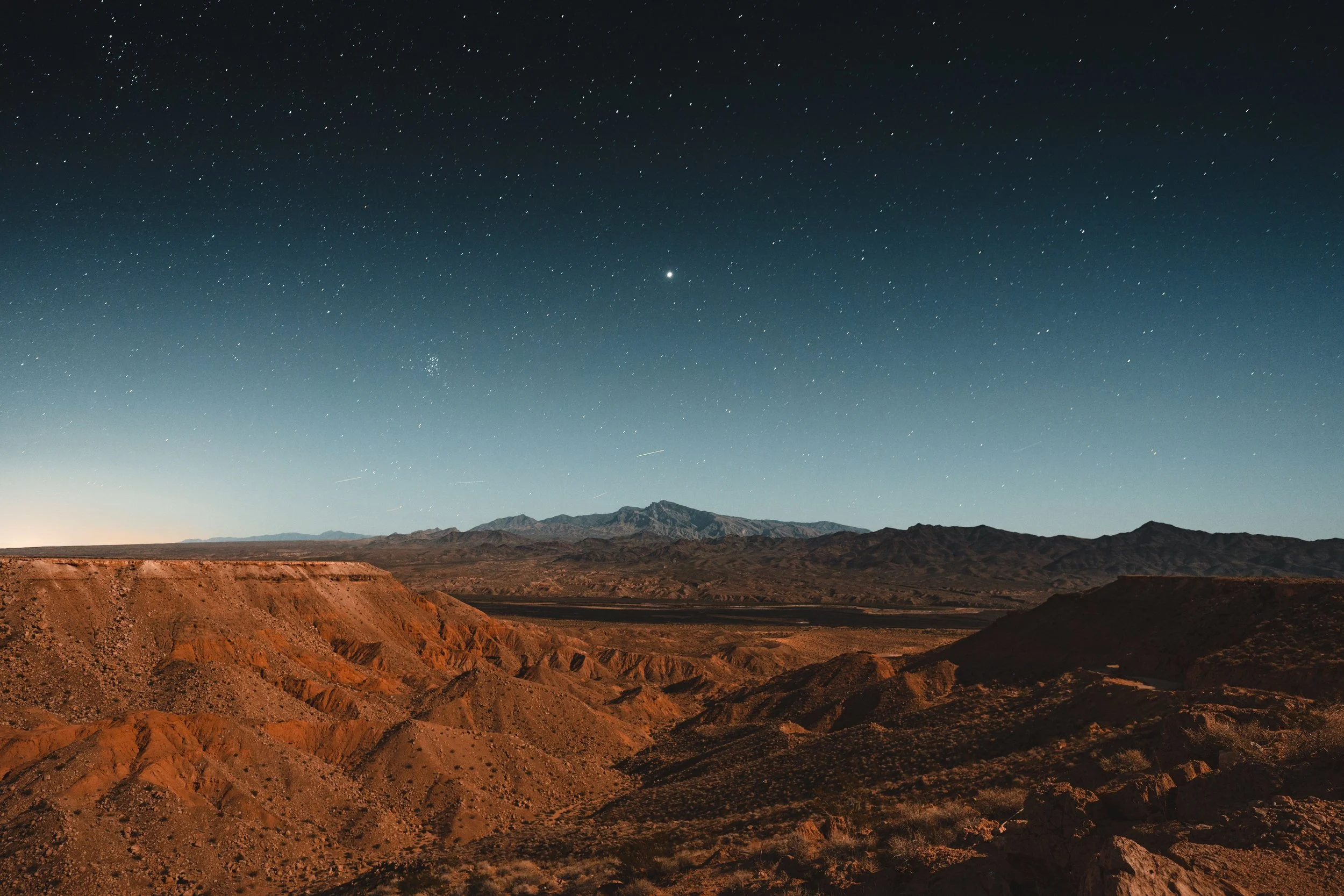 Nighttime desert landscape with rugged, reddish-brown hills under a starry sky with a bright planet or star visible.