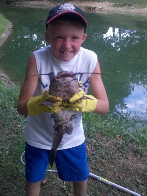 A young boy smiling and holding a large catfish by the water's edge, wearing a baseball cap, a sleeveless shirt, yellow gloves, and blue shorts.