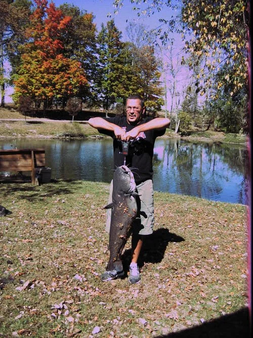 A man stands on grass near a pond, holding a large catfish with both hands. The background features trees with colorful fall foliage and a bright, partly cloudy sky.