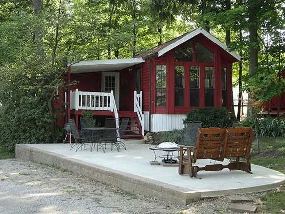 Cabin with large front porch overlooking a pond, surrounded by trees, outdoor patio with metal table and chairs, a wooden bench, and a small fire pit.