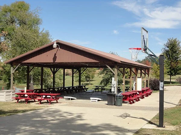 A park pavilion with a basketball hoop nearby and several picnic tables under the shelter, surrounded by trees and a clear sky.