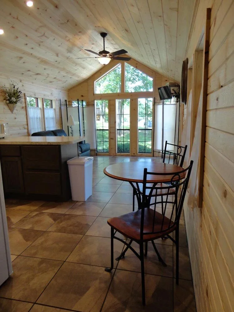 View of a cozy wooden cabin interior with large windows, a ceiling fan, a small dining table with chairs, a kitchen counter, and a TV mounted on the wall.