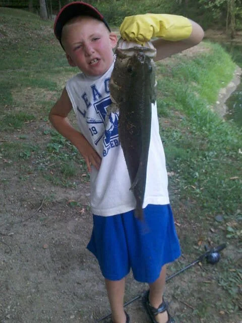 A young boy holding a large fish he caught, standing outdoors near a water body.