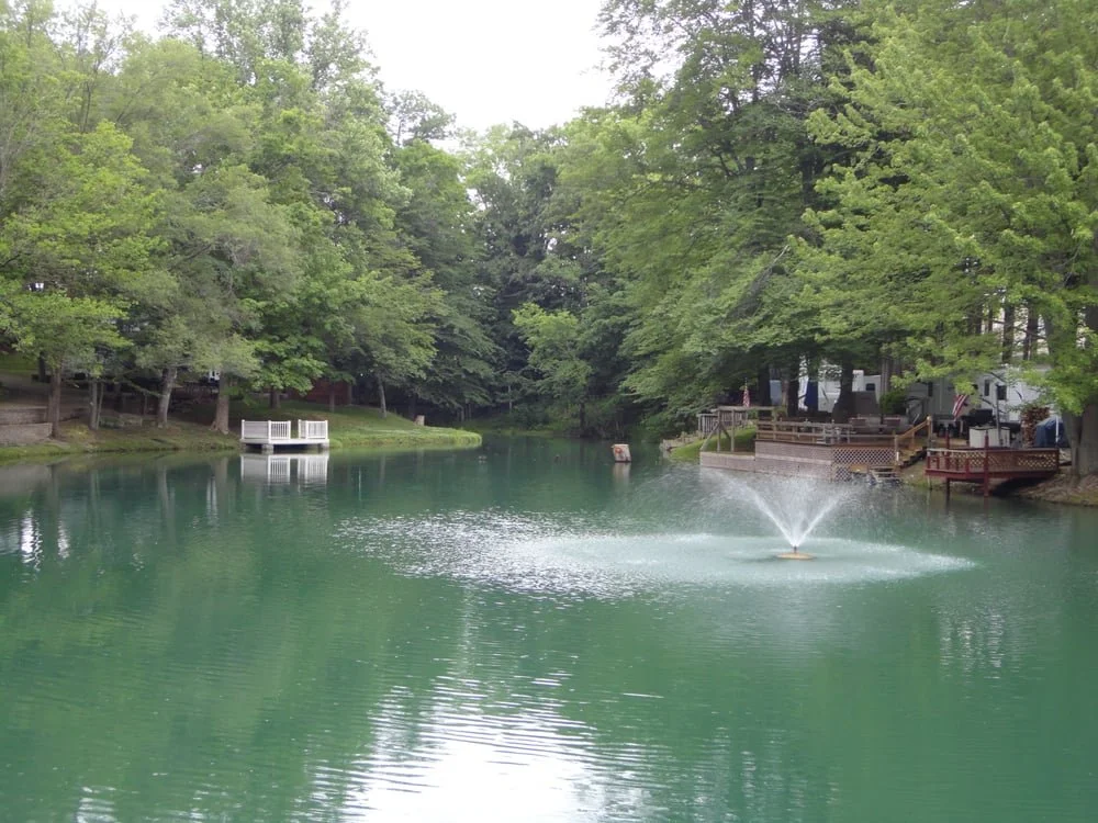 A peaceful pond surrounded by green trees with a fountain in the middle and a housing area with decks along the shoreline.