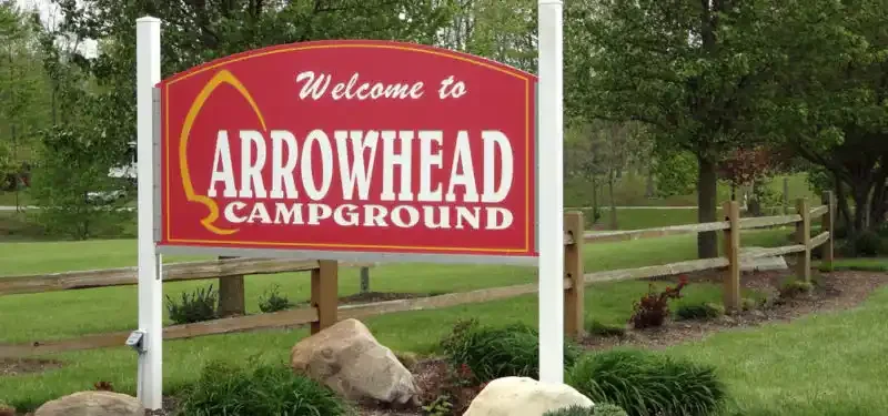 Welcome sign for Arrowhead Campground, with a red background, white text, and an arrow graphic, set beside grassy area with rocks and trees.