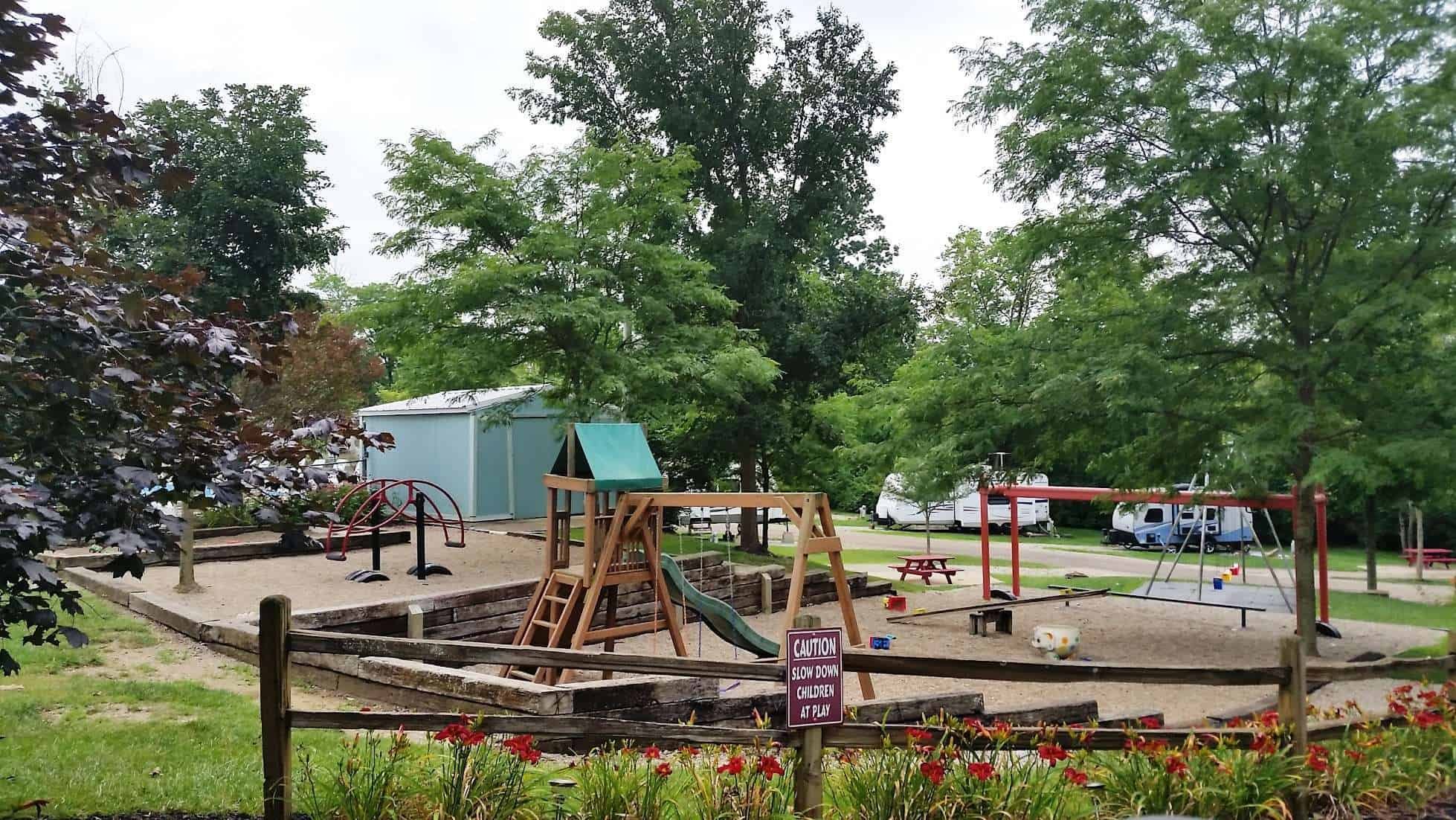 A playground with a slide, swings, and a sandbox area surrounded by trees and a fence, with RVs in the background and a caution sign at the front.