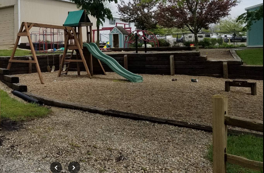 Childrens playground with a wooden swing set, slide, sandbox, and various playground equipment surrounded by wood edging, trees, and a fence in background.