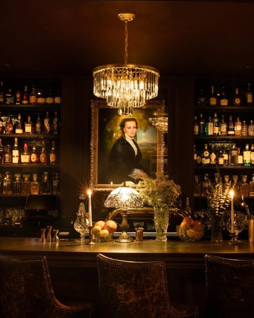 Elegant bar interior with a large chandelier, shelves of liquor bottles, a portrait of a woman, and lit candles on the bar counter.