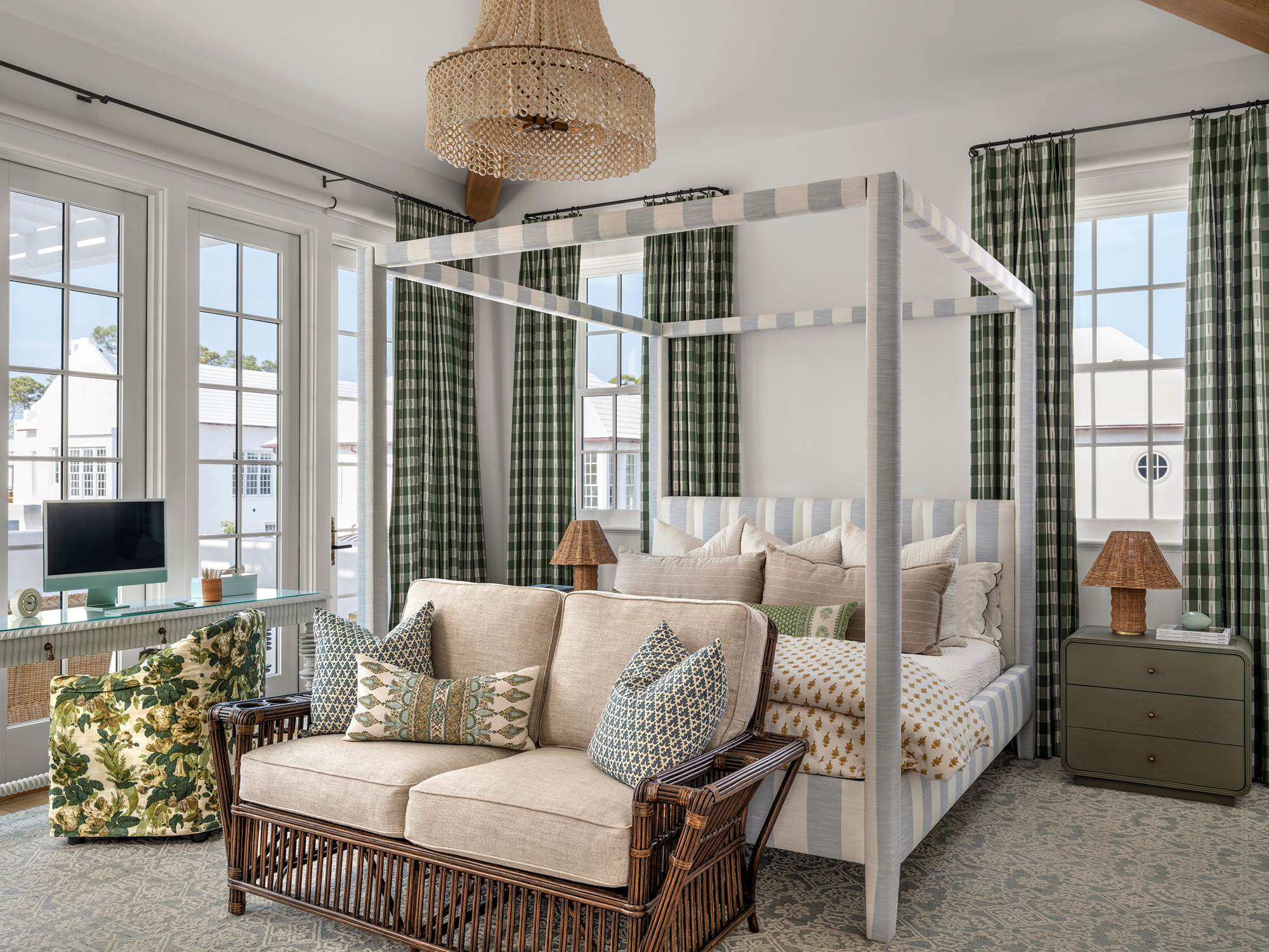 A bedroom with large windows, a four-poster bed with striped fabric, a green nightstand with a wicker lamp, a beige sofa with patterned pillows, and a wicker ottoman in front of it. There are green checkered curtains and a woven chandelier hanging fr