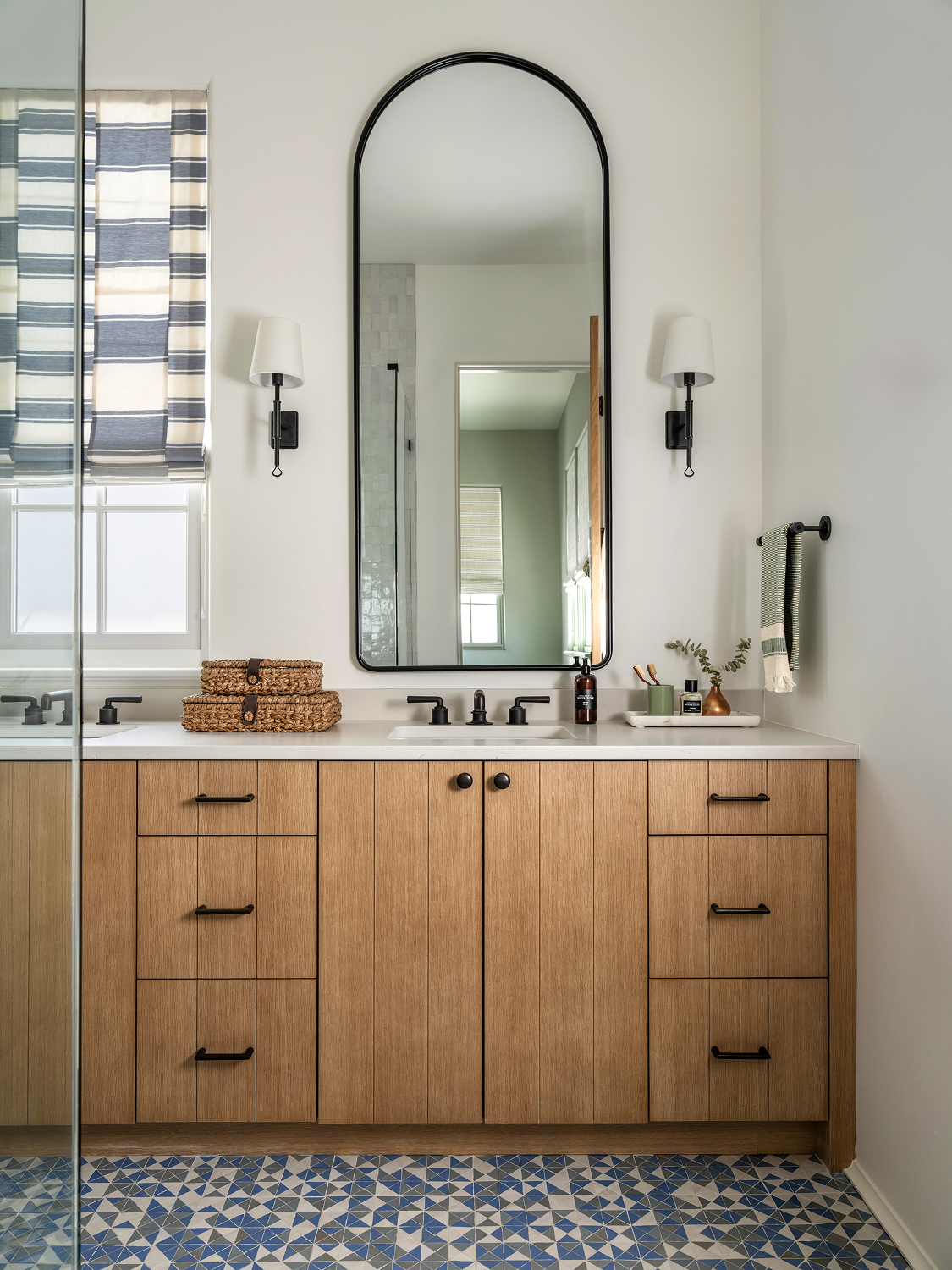 Bathroom vanity with wooden cabinets, white countertop, and oval mirror, flanked by wall-mounted lamps, decorated with baskets, soap dispenser, and small plant, with a window and patterned tile floor.