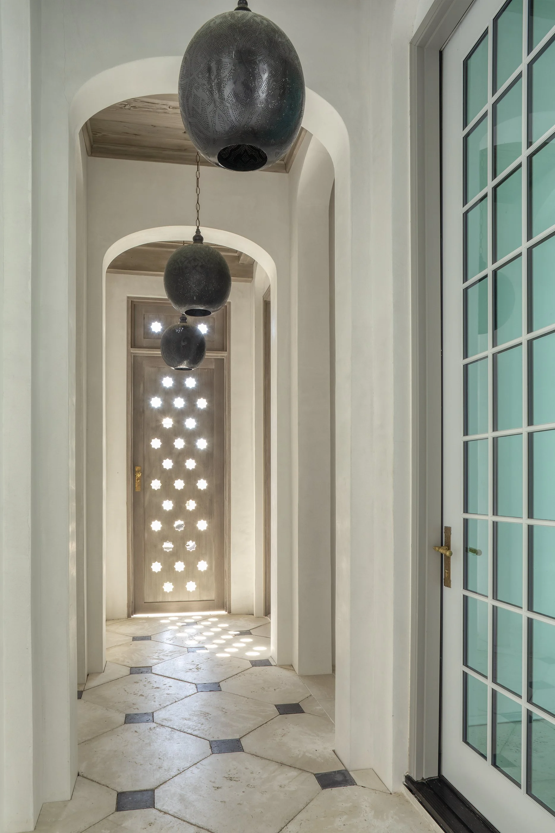A hallway with arched openings, hanging black pendant lights, a decorative door with star-shaped cutouts, and a floor with large hexagonal tiles and smaller dark square tiles.