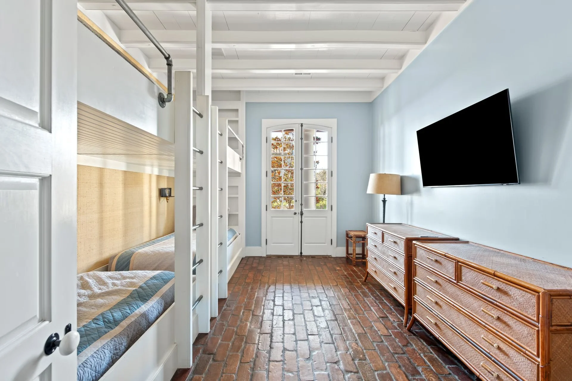 Interior of a living space with brick flooring, a wall-mounted flat-screen TV, a wooden dresser, and a window with French doors opening outside.
