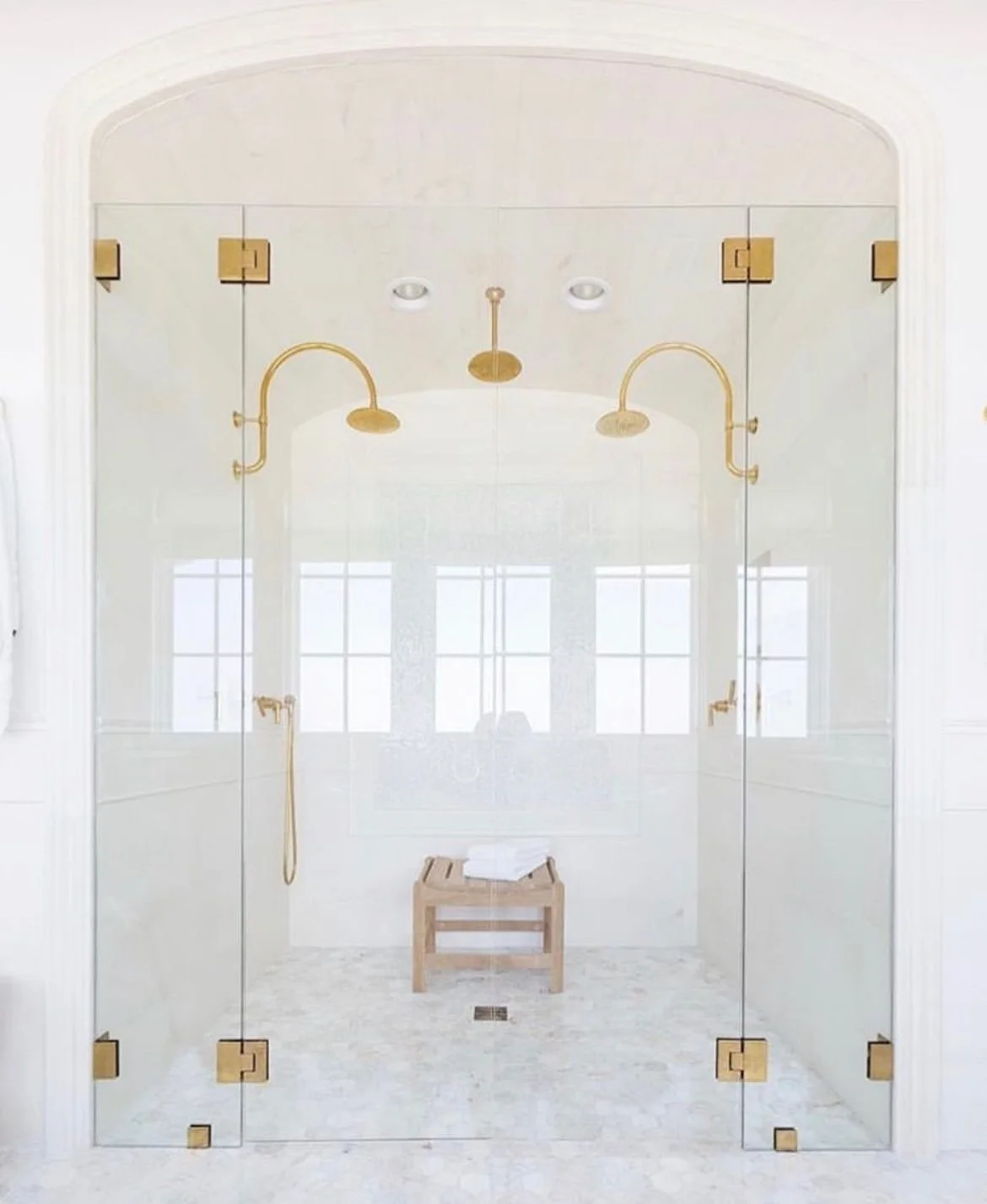 A luxurious walk-in shower with glass walls, two gold showerheads, a wooden stool with white towels, and large windows allowing natural light.