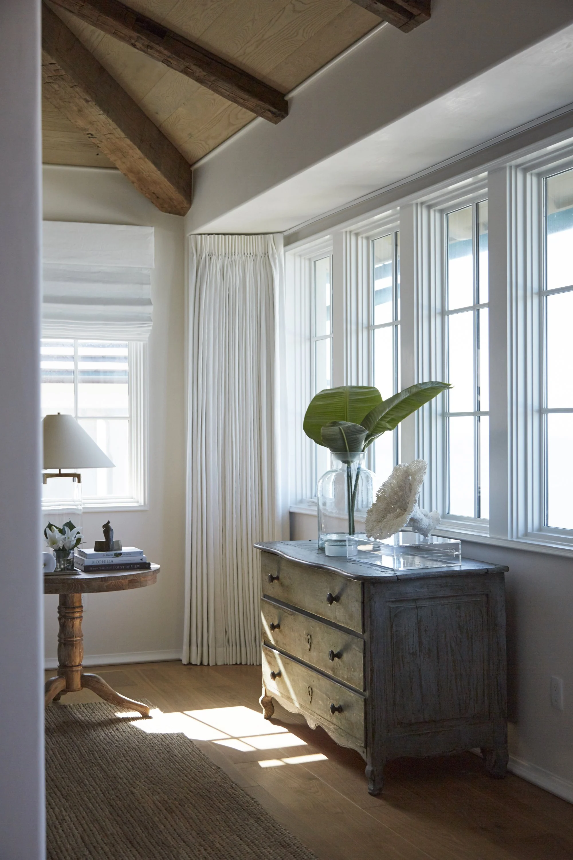 Sunlit corner of a room with a vintage wooden dresser holding a glass vase with large green leaves and a white decorative object, cream curtains, a wooden side table with books, a small sculpture, and a lamp, along with hardwood flooring and a rug.