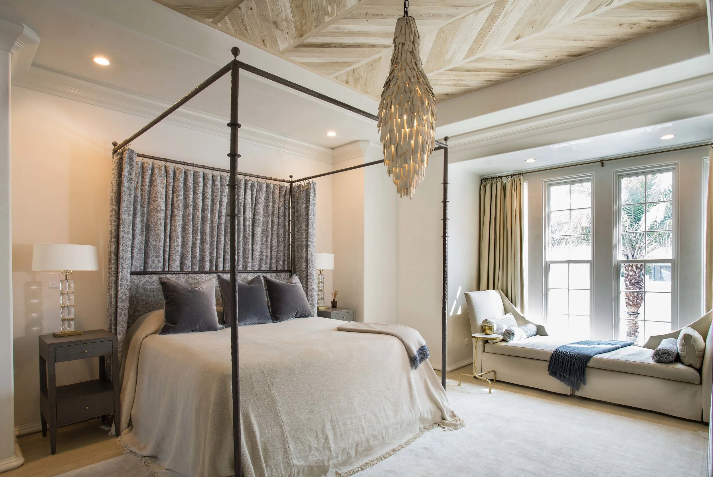 A bedroom with a four-poster bed featuring dark metal frame and draped curtains, cream bedding with gray pillows,-side table with a lamp, and a window with beige curtains and a chaise lounge by the window with towels and a sweater draped over it.