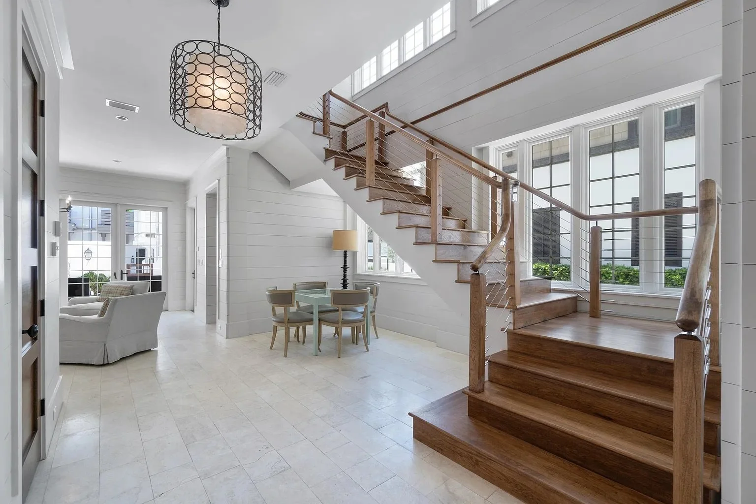 Bright open living space with white walls, wooden stairs, large windows, a small dining table with four chairs, a beige armchair, and a round chandelier.