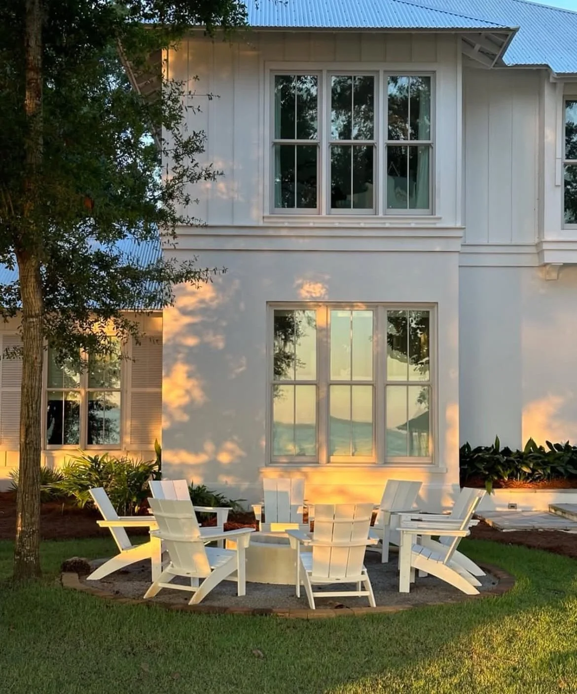White house with multiple large windows, surrounded by green grass, shrubs, and trees. In front, there is a circular arrangement of white outdoor chairs around a fire pit, with a sunset casting warm light and shadows on the house and yard.