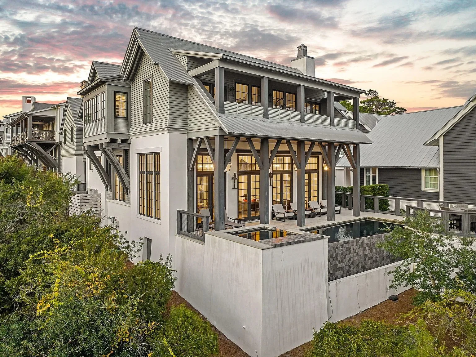 Modern multi-story house with large windows, a rooftop deck, and outdoor lounge area with a hot tub, set against a sunset sky.