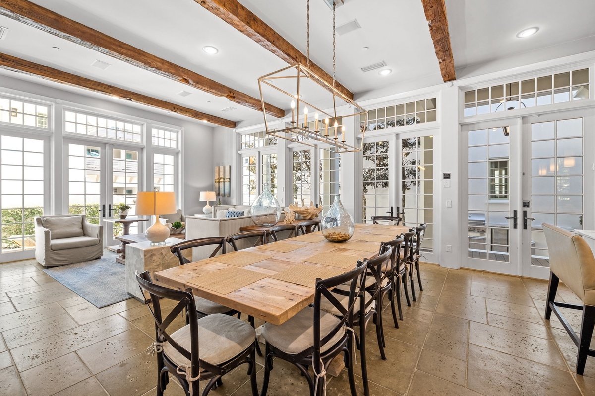 Bright dining and living area with large windows, exposed wooden ceiling beams, a wooden dining table with twelve chairs, and a cozy seating area with armchairs and sofas.