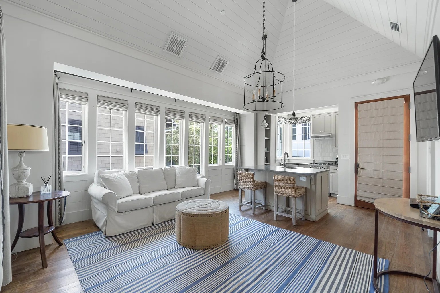 Cozy open-concept living room and kitchen with white walls, large windows, beige sofa, striped blue rug, wooden furniture, and kitchen island with bar stools.
