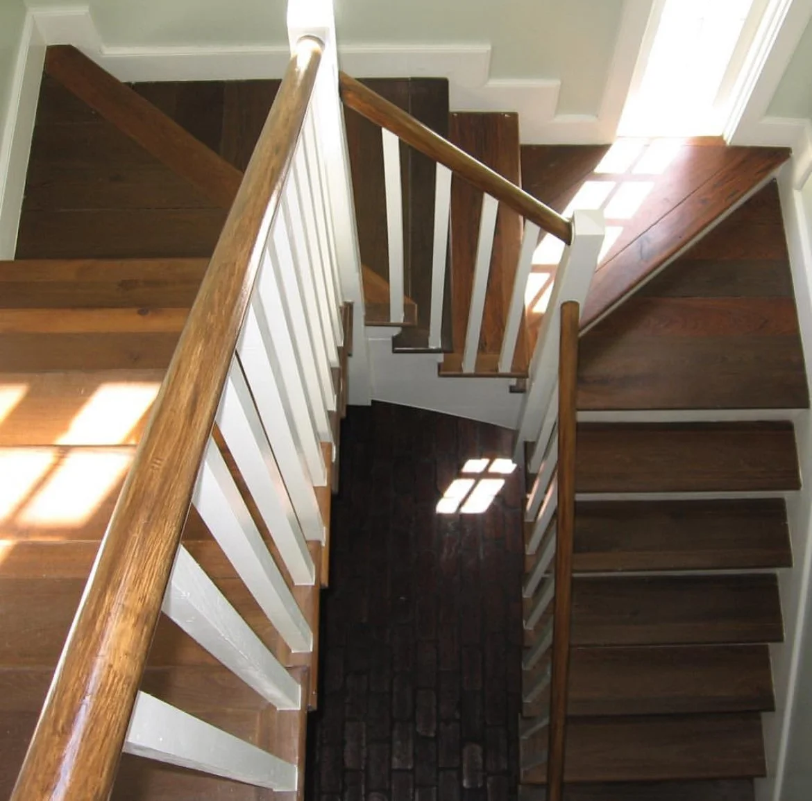 Top-down view of a staircase with dark wooden steps, white risers, and wooden handrails, with sunlight creating shadows on the stairs and a brick-patterned mat on the floor at the bottom.