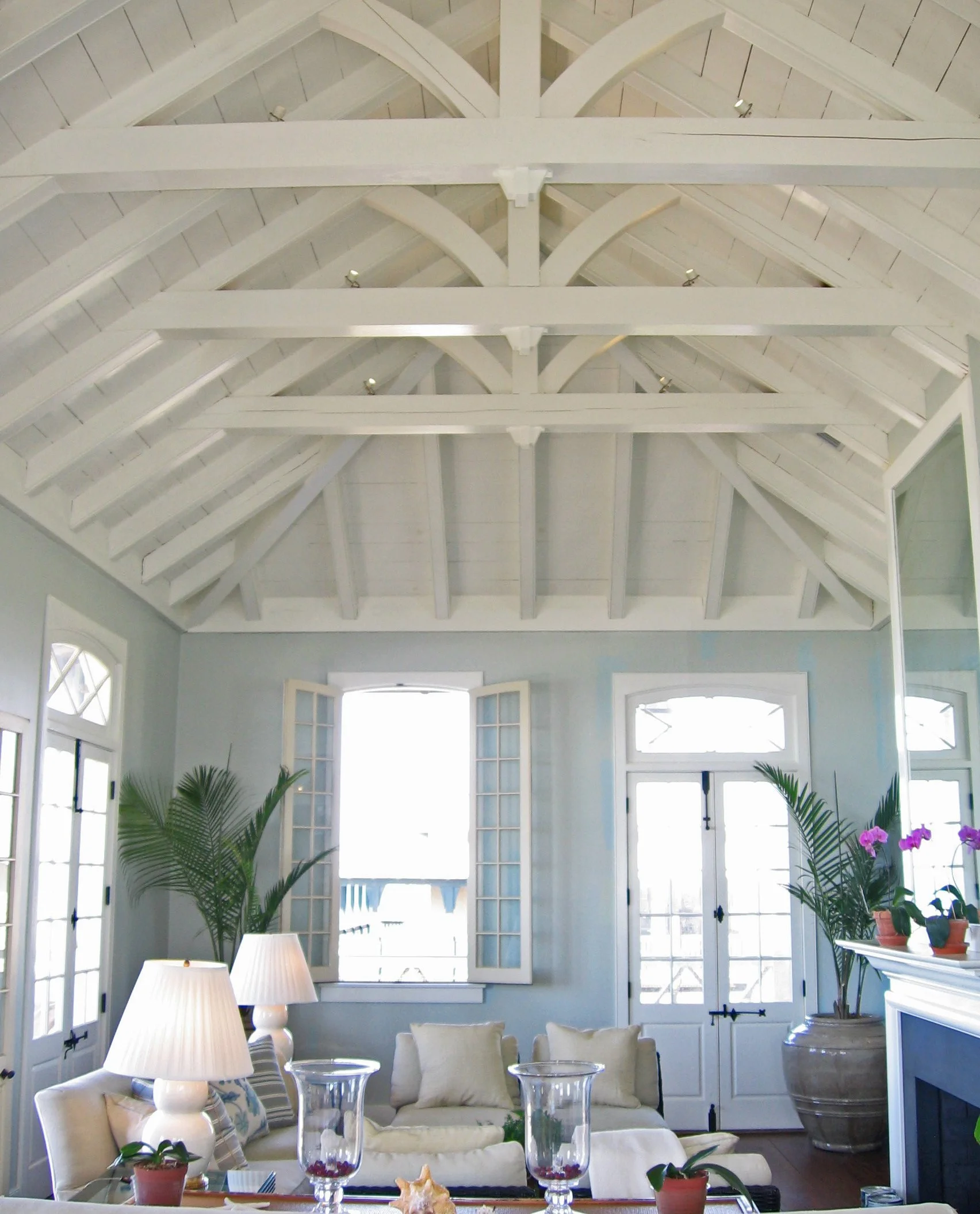 Living room with high vaulted white ceiling, large windows with open shutters, potted plants, sofa with pillows, lamps, and decorative items.
