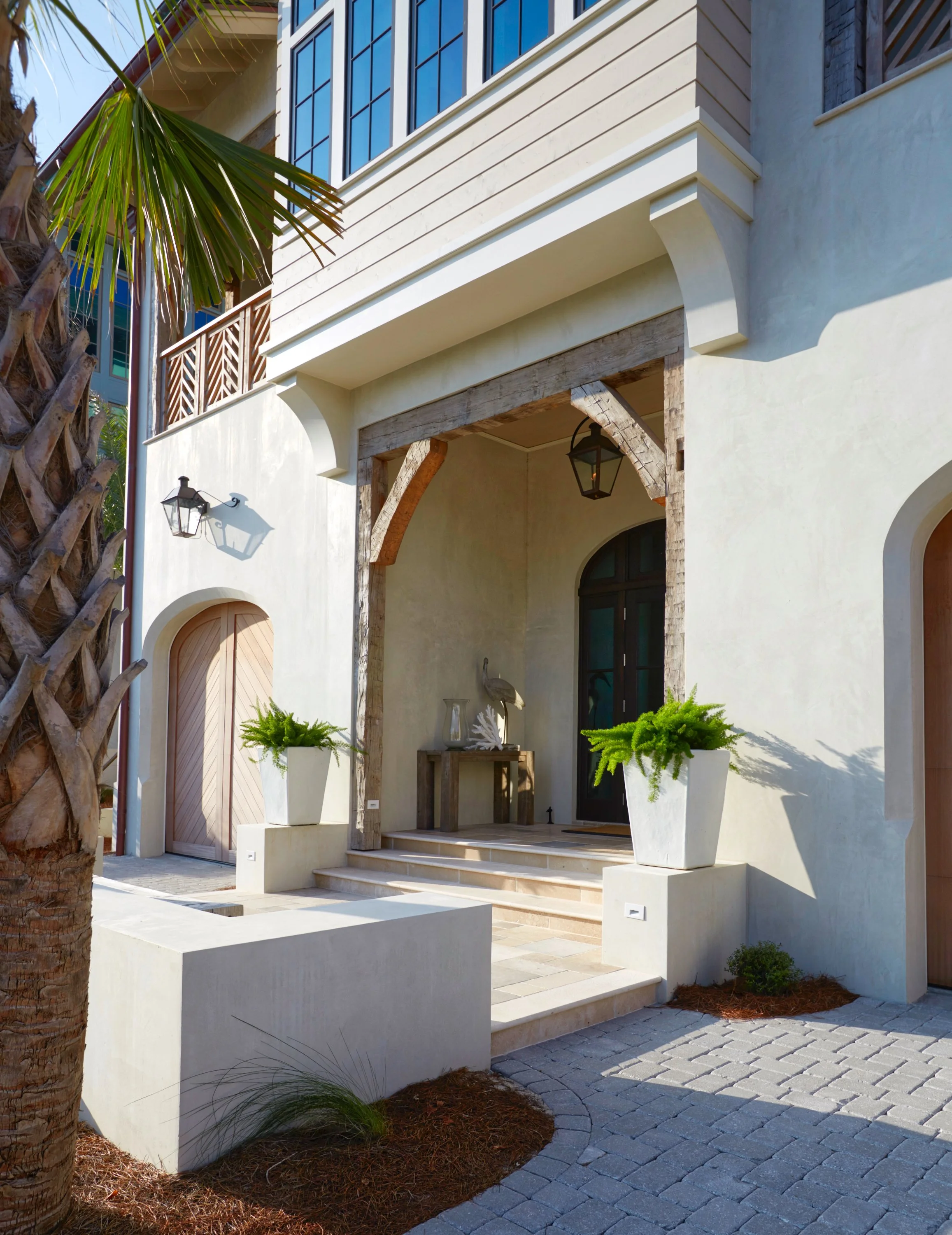 Front view of a modern house with a beige exterior, stone accents, and large windows, featuring a porch with steps and potted green plants, under a sunny sky.