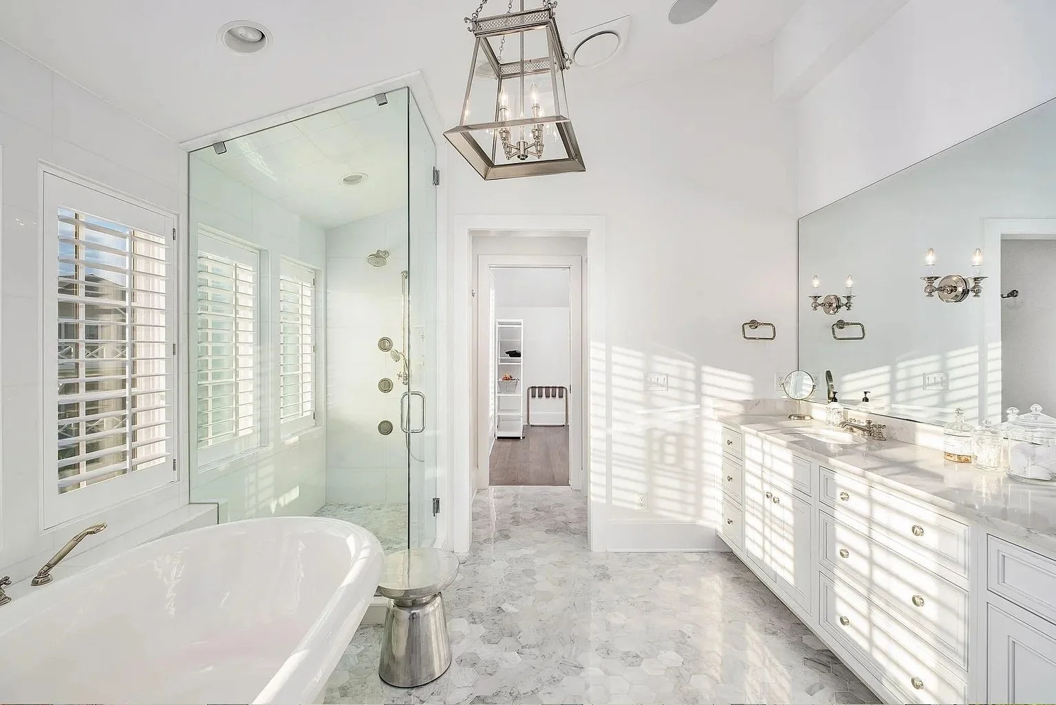 Bright, white bathroom with a freestanding bathtub, double vanity with large mirror, walk-in shower with glass door, and a separate room with a toilet. Shutters on the windows cast striped shadows on the walls and floor.