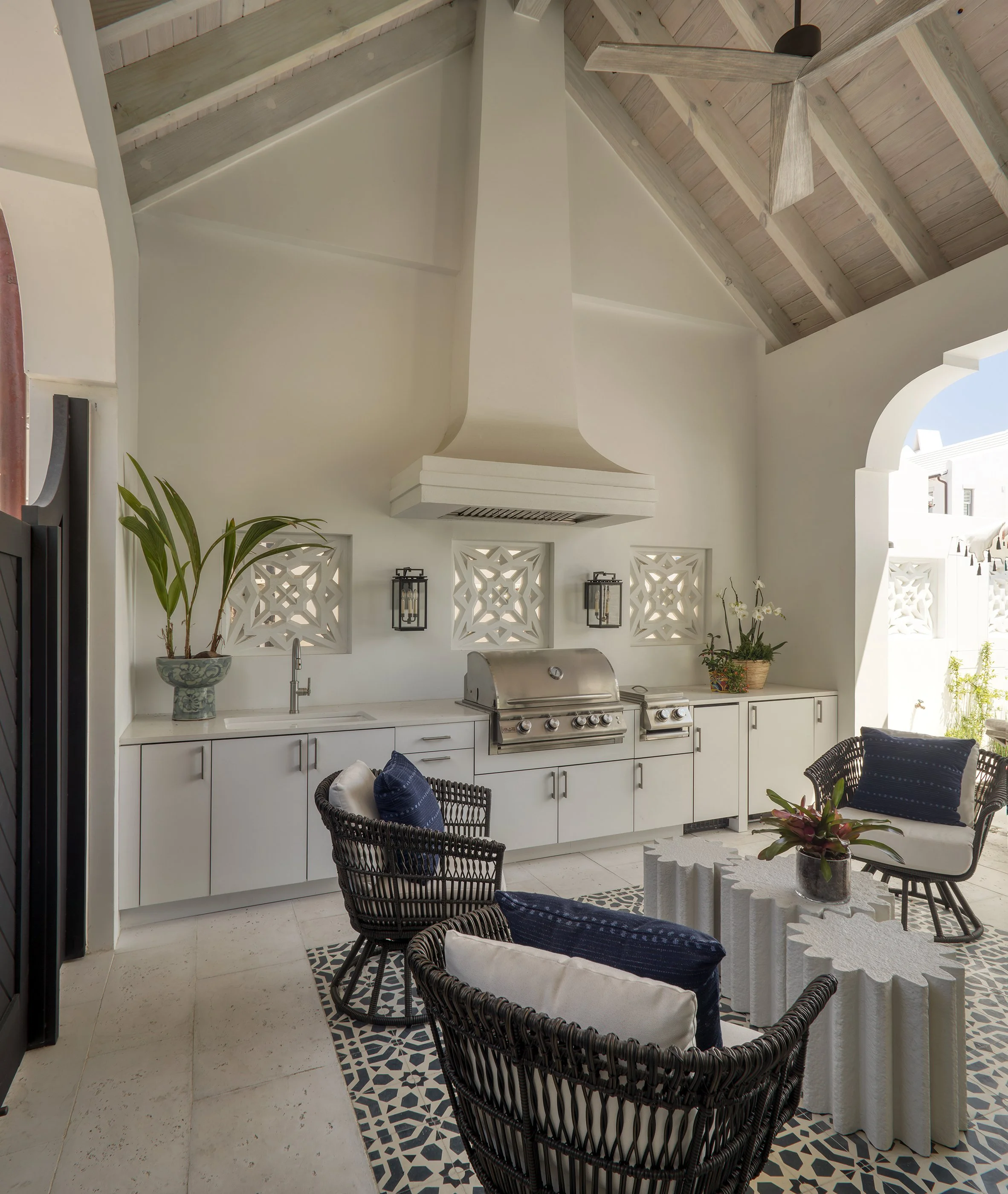 Outdoor patio area with a built-in grill, seating with black wicker chairs and white cushions, and a decorative white table. There are potted plants on the counter, patterned tile flooring, and lantern-style wall lights. The space has a vaulted ceili